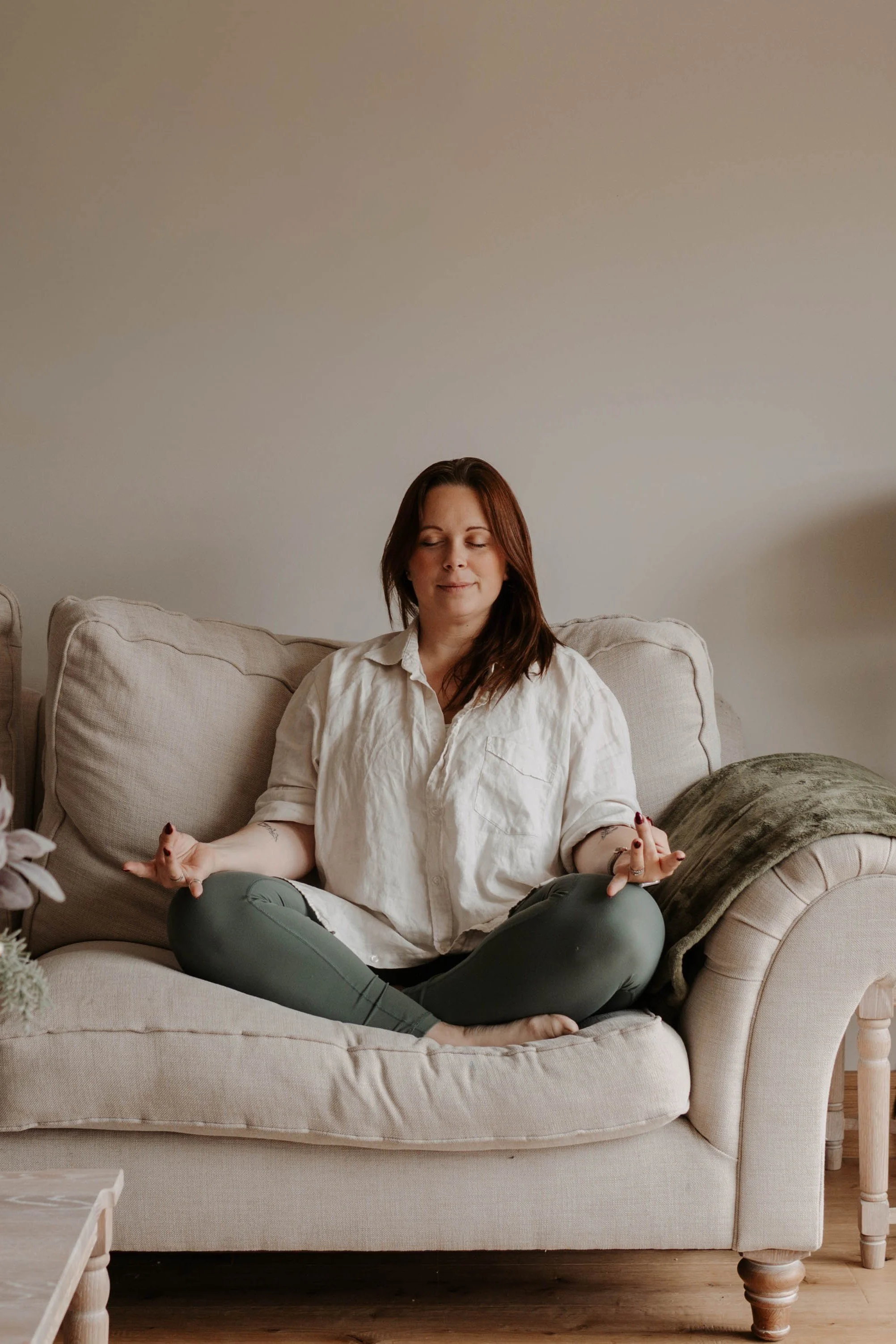 A woman practicing meditation on a beige sofa at home with her eyes closed and a peaceful expression.