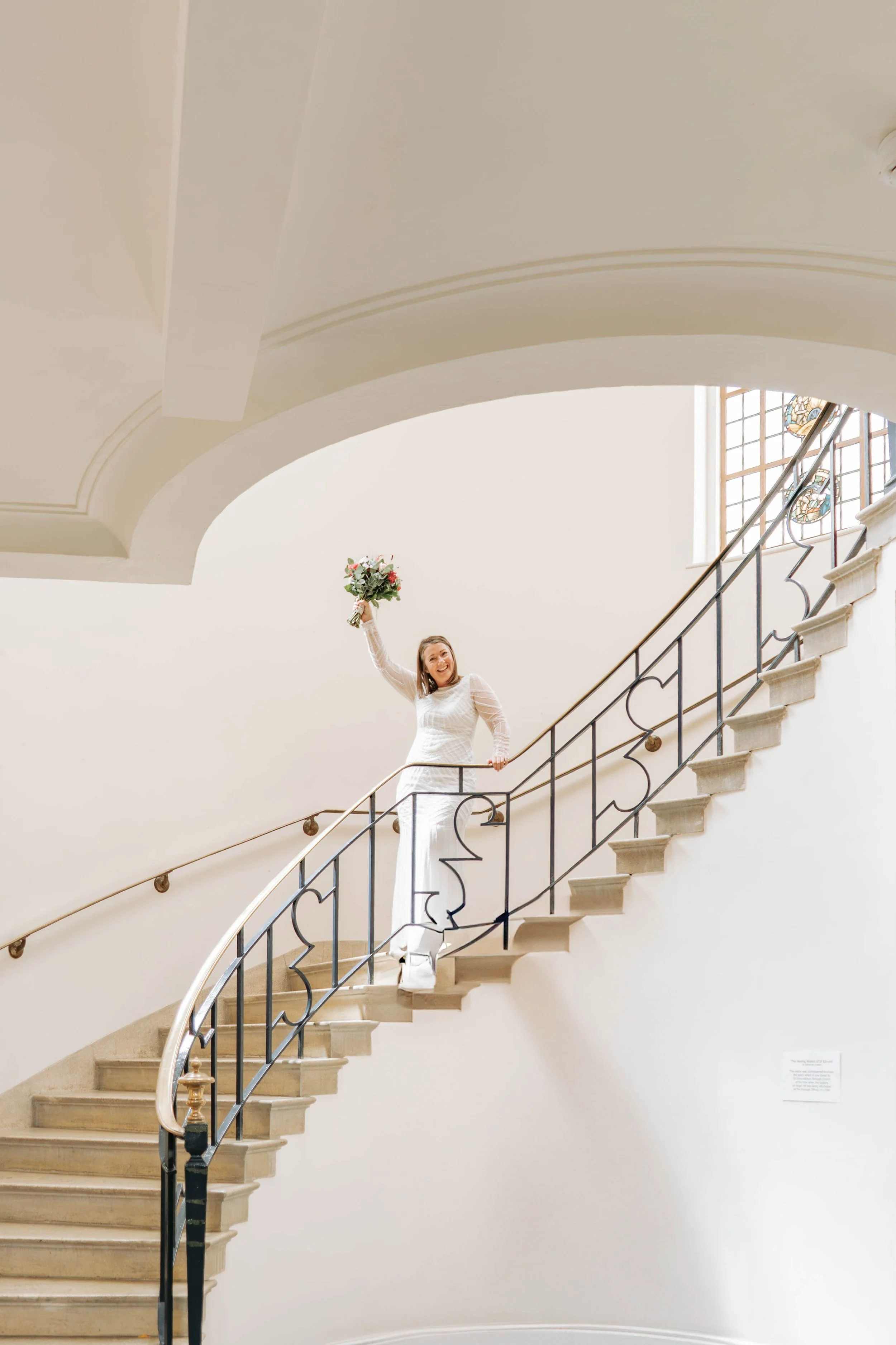 A woman in a white wedding dress holding a bouquet of flowers, standing on a curved staircase with black iron railing, inside a building with white walls and a stained glass window.