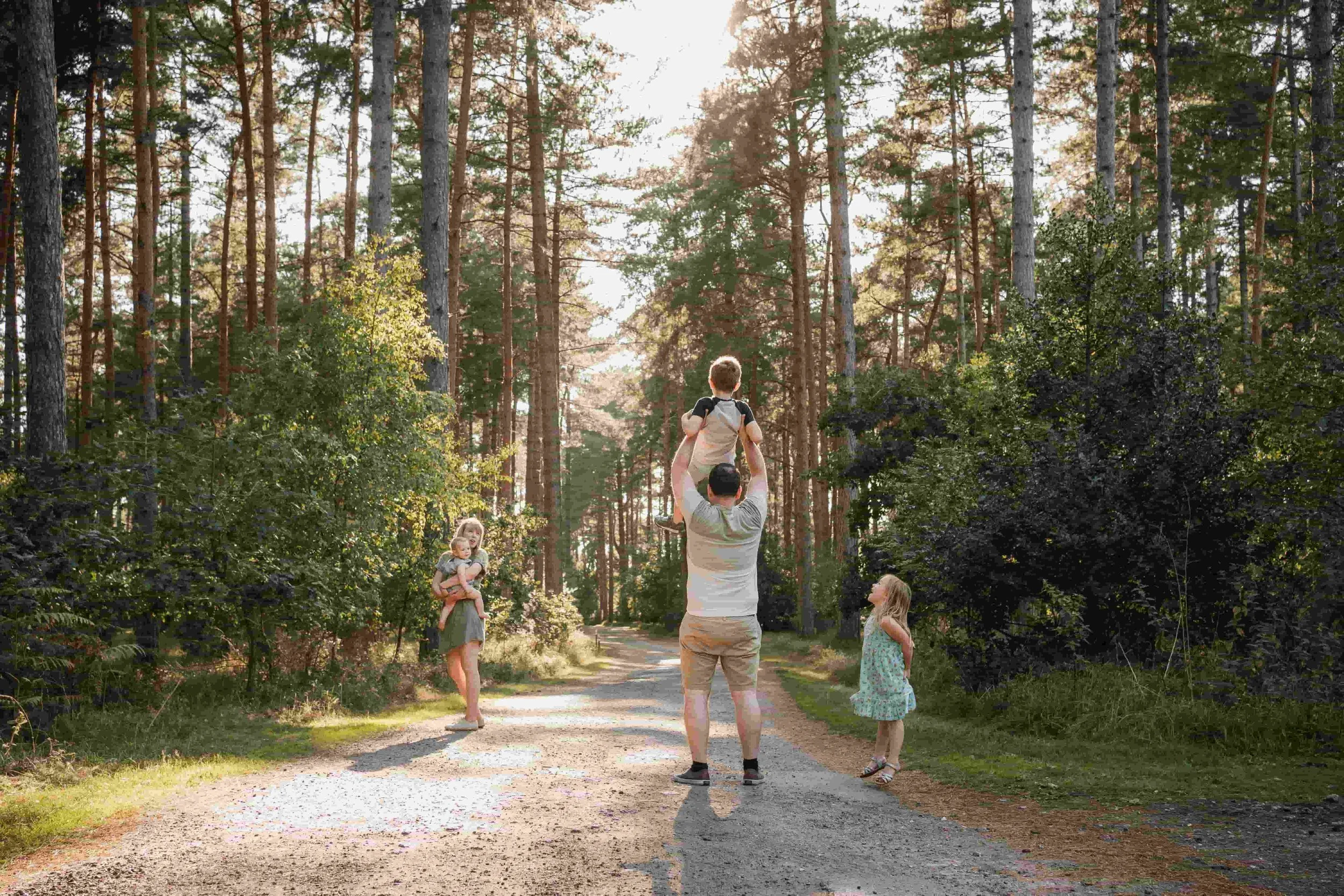 A man is holding children on his shoulders while they walk along a dirt trail in a forest, with two young girls standing nearby and tall trees surrounding them.