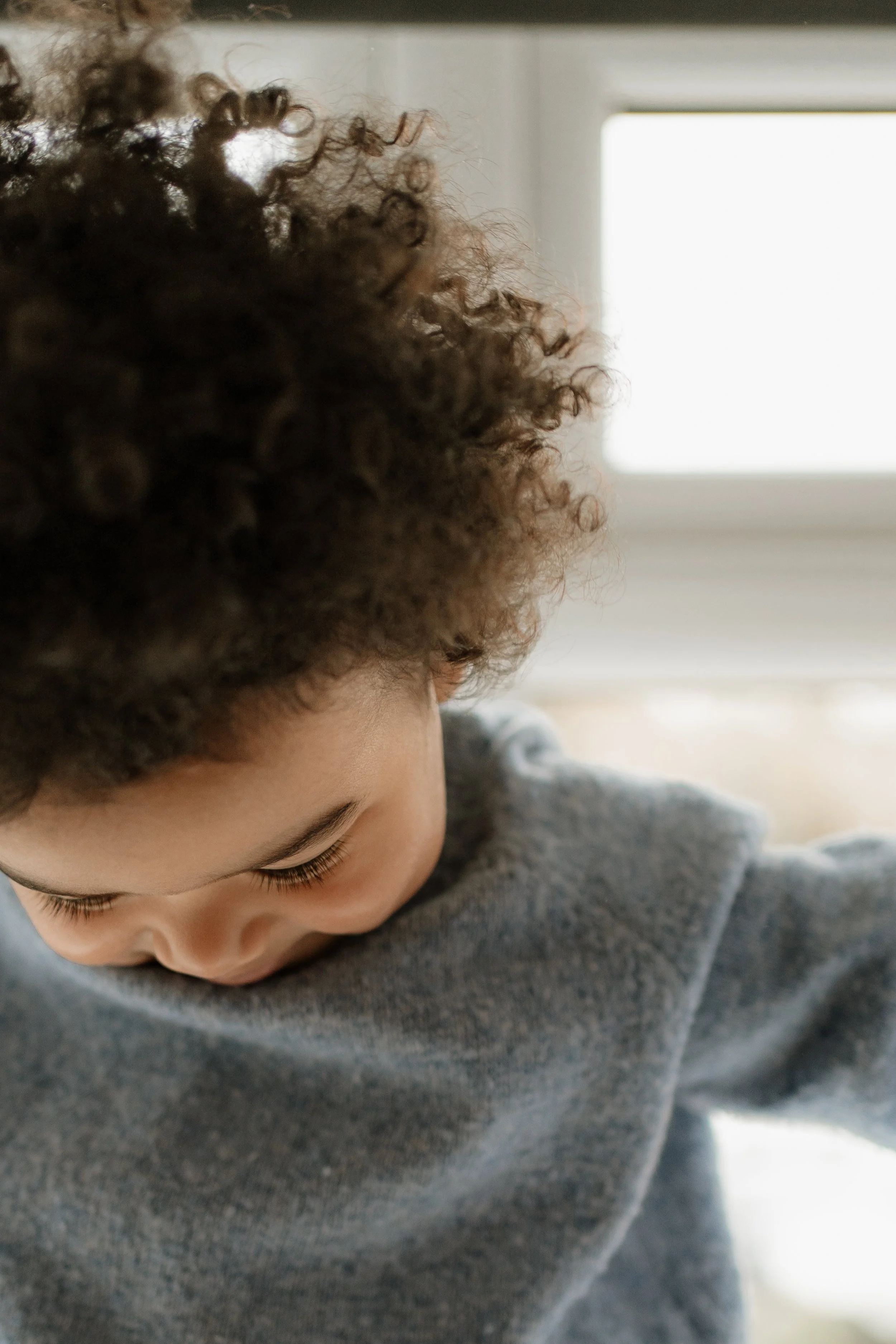 Close-up of a young child with curly hair, looking down at something outside a window.