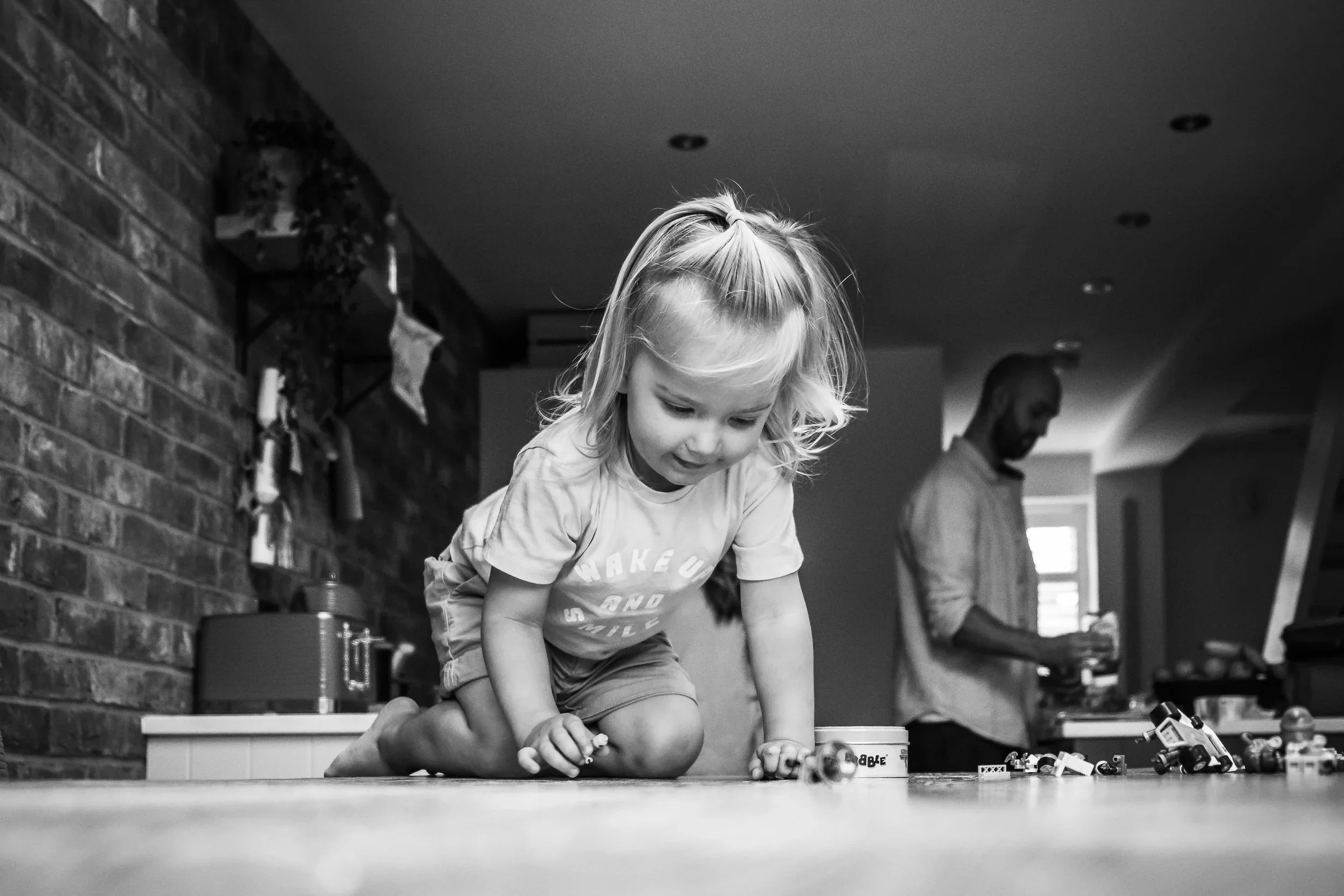 A young girl with blonde hair playing on the floor with toys, while a man stands in the background in a cozy home setting.
