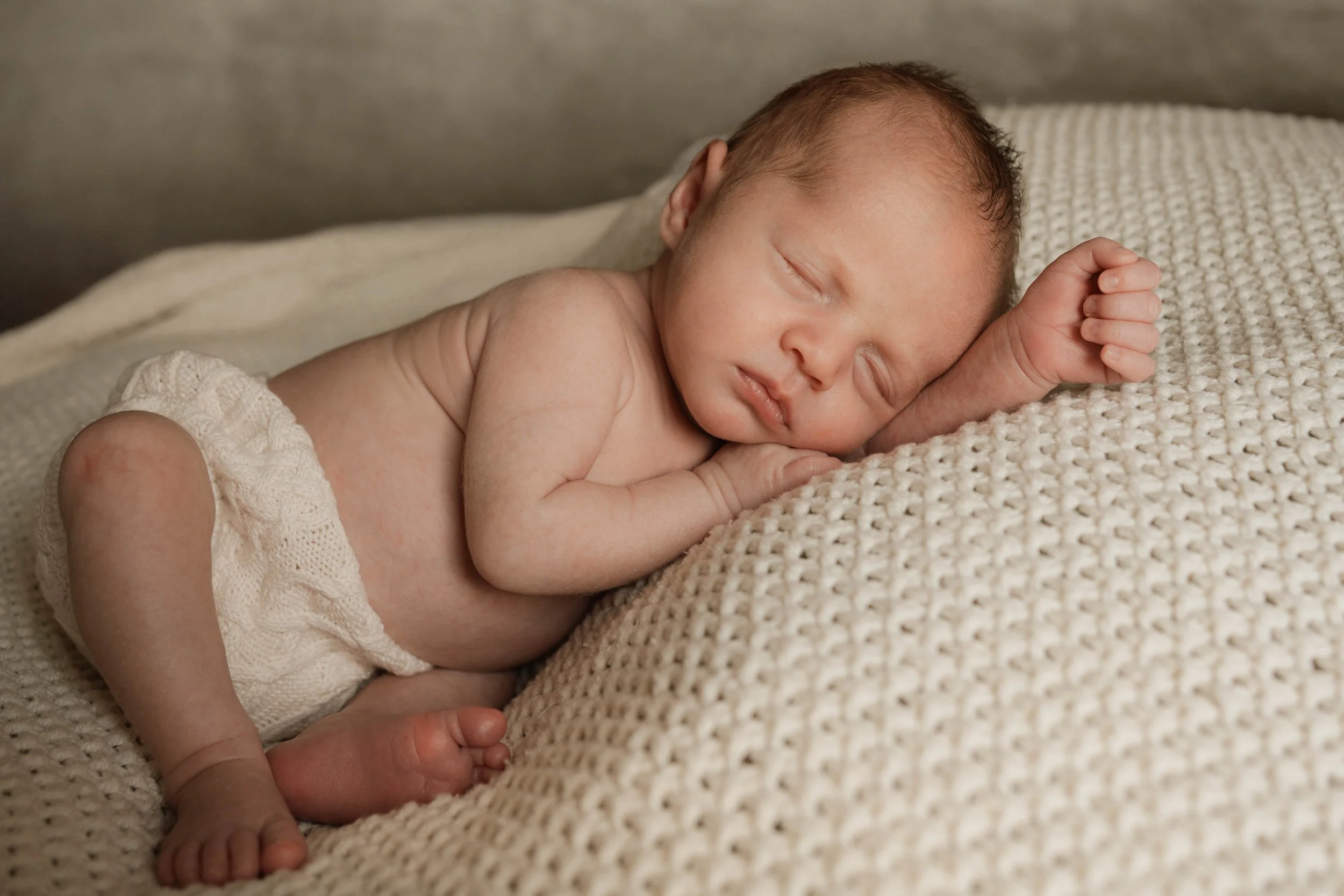 A sleeping newborn baby lying on a soft, textured cream-colored blanket, with a beige sweater on, peacefully resting with eyes closed and arm bent at the elbow.