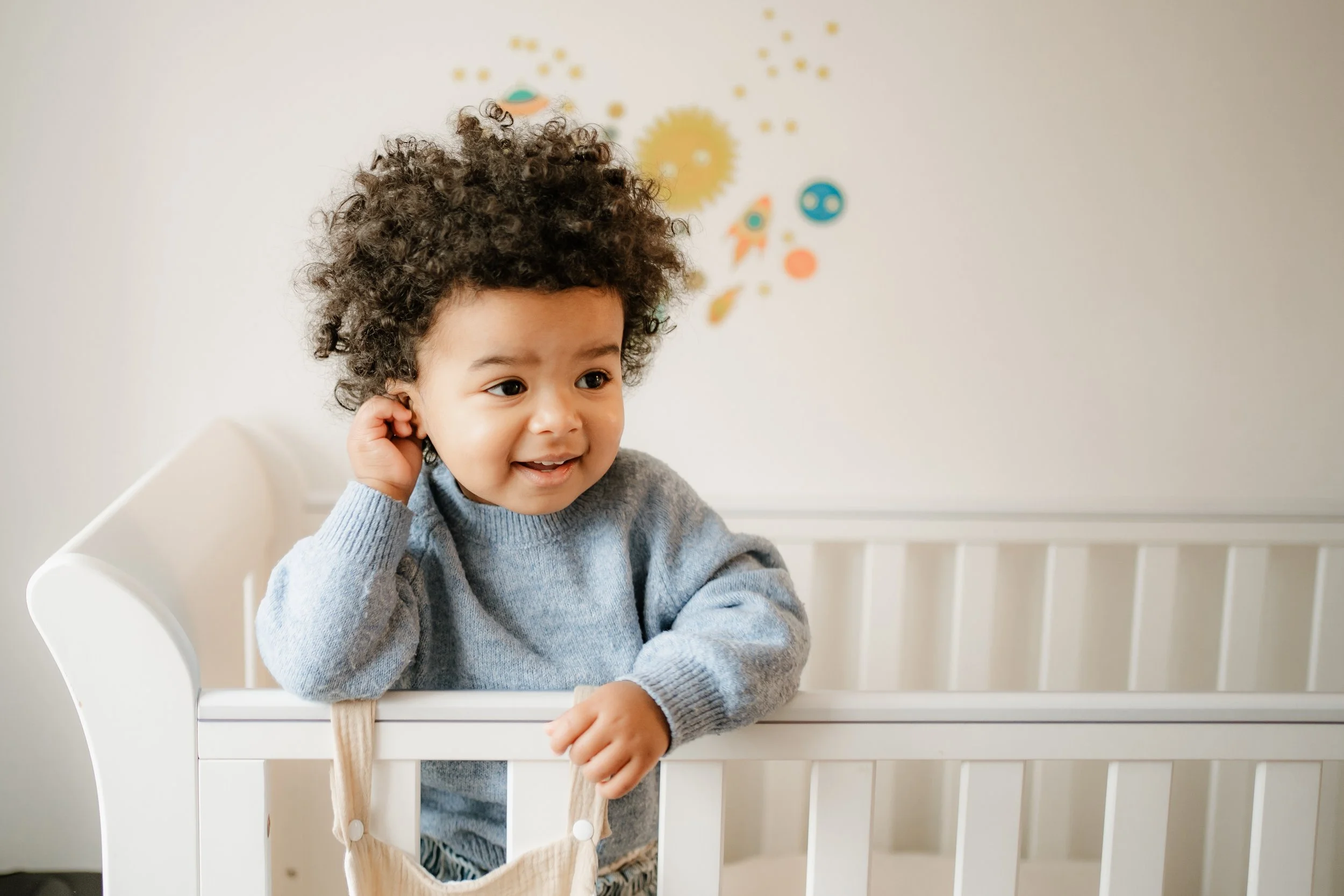 A young child with curly hair standing in a white crib, smiling and touching their ear, wearing a grey sweater. There are colorful wall stickers behind in a space theme.