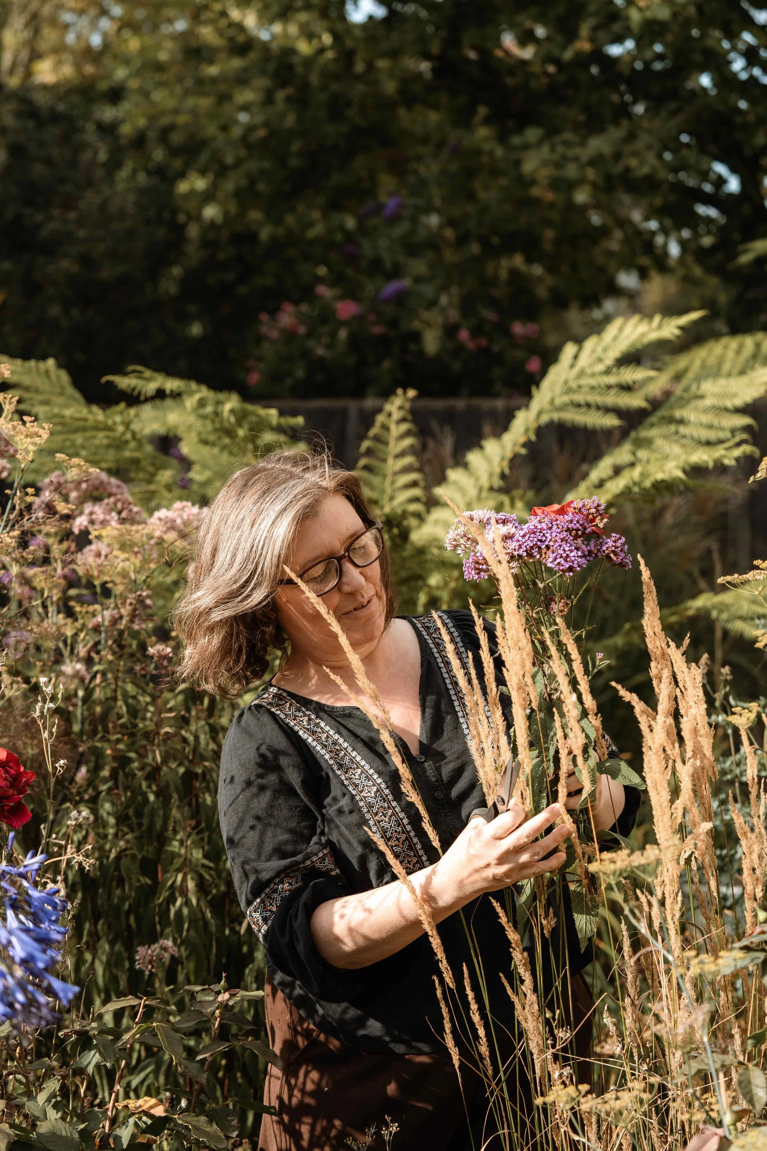 A woman with glasses, shoulder-length hair, wearing a black embroidered blouse, is outdoors in a garden surrounded by various flowers and tall grasses, holding a plant in her hands.