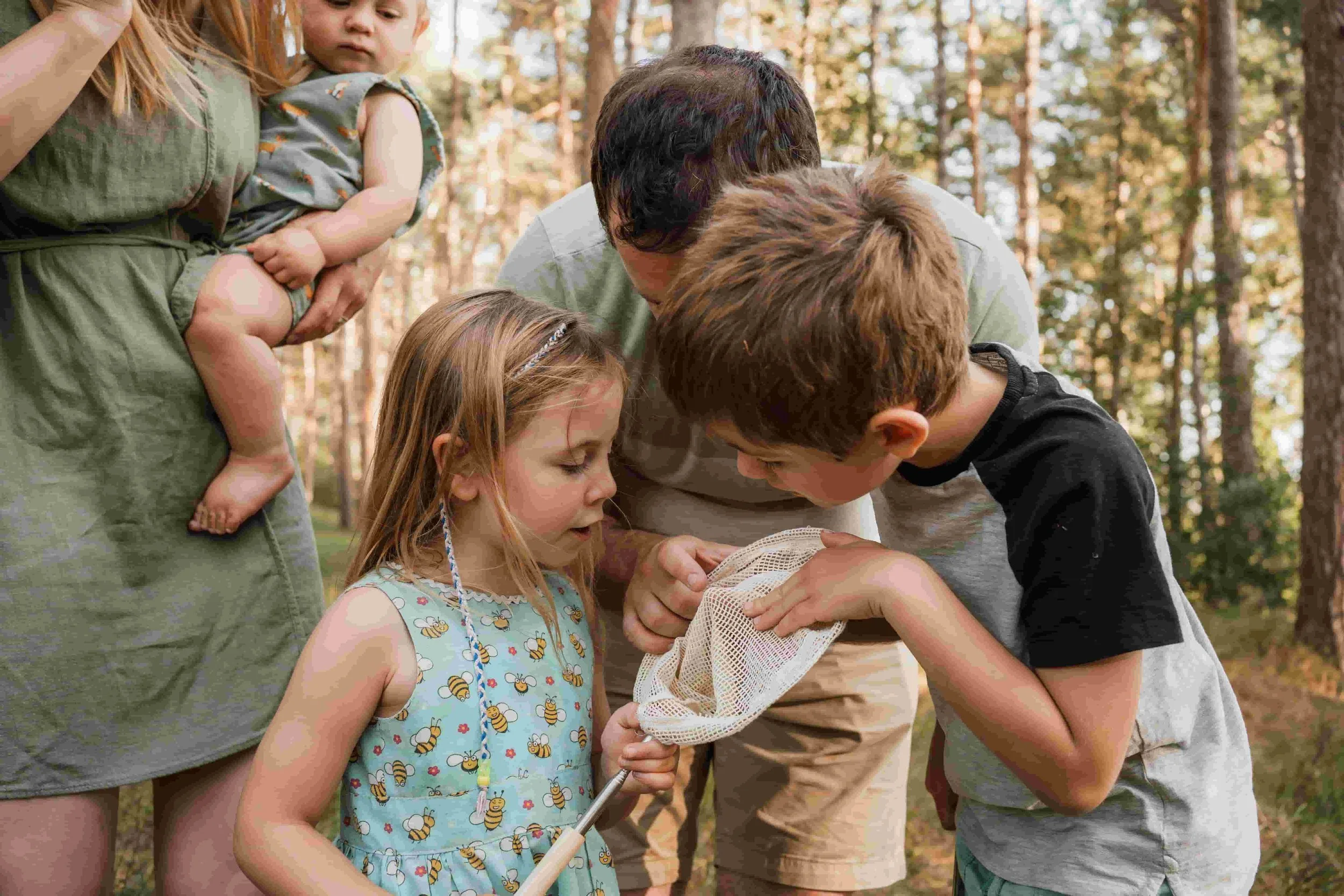Children and adults in a forest gathered around, looking at a small animal or object in a net held by one of the children.