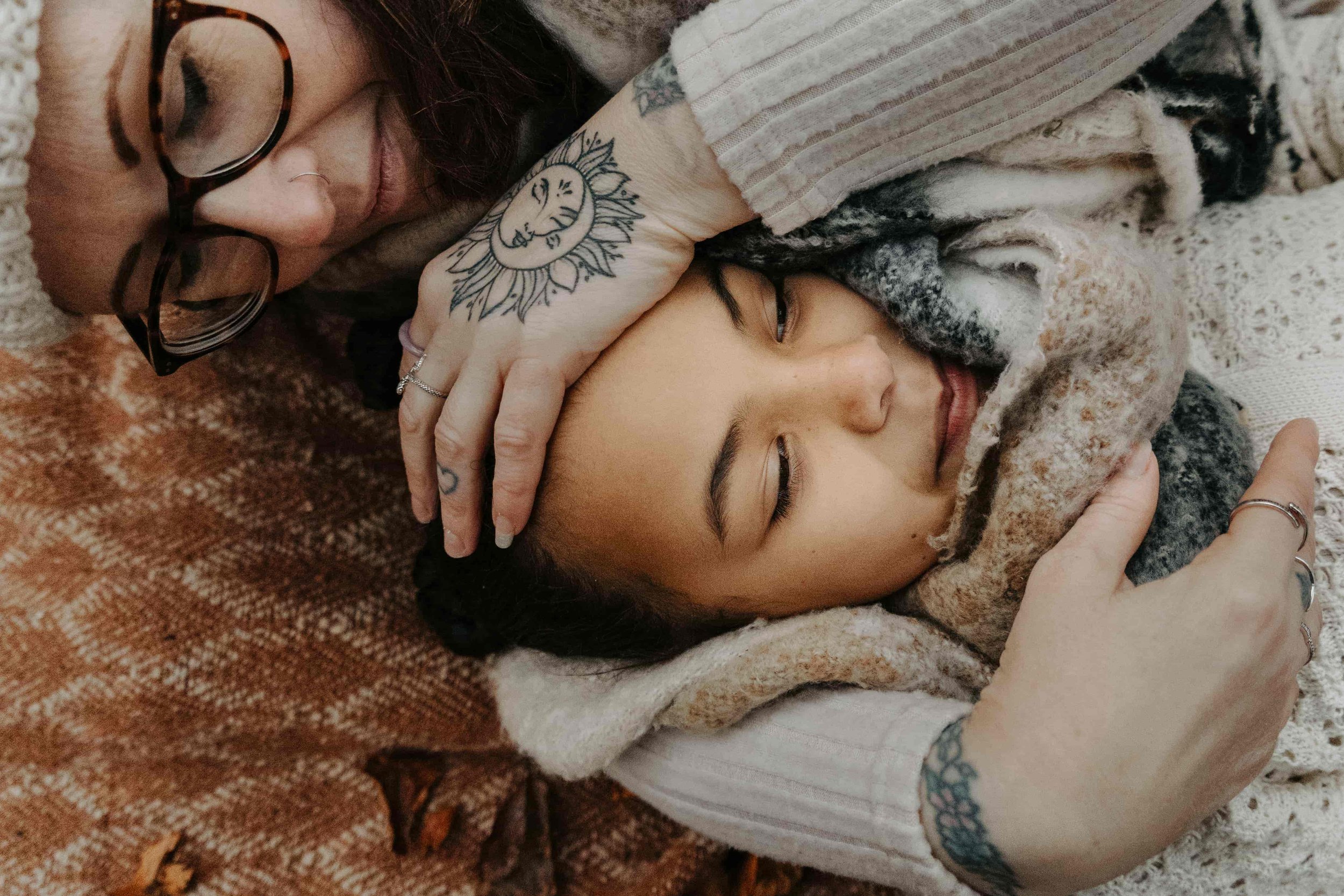 A woman with glasses and tattoos gently embraces a young boy on a knit blanket, suggesting an intimate and tender moment.