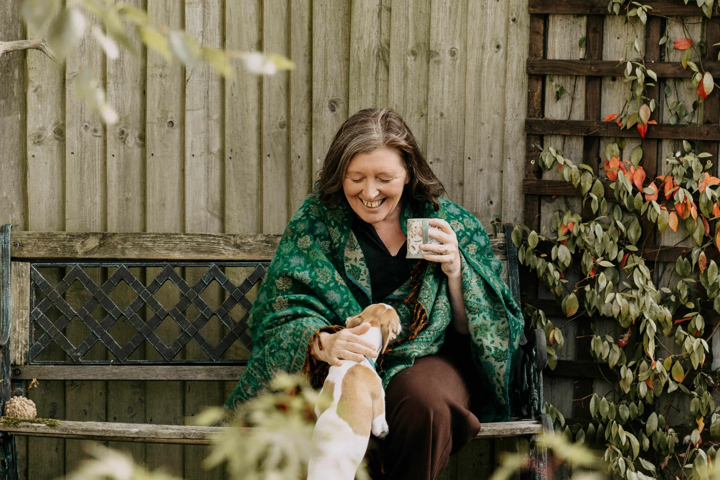 A smiling woman with gray hair in a green shawl sitting on a wooden bench, holding a small puppy, in a backyard with a wooden fence and foliage.