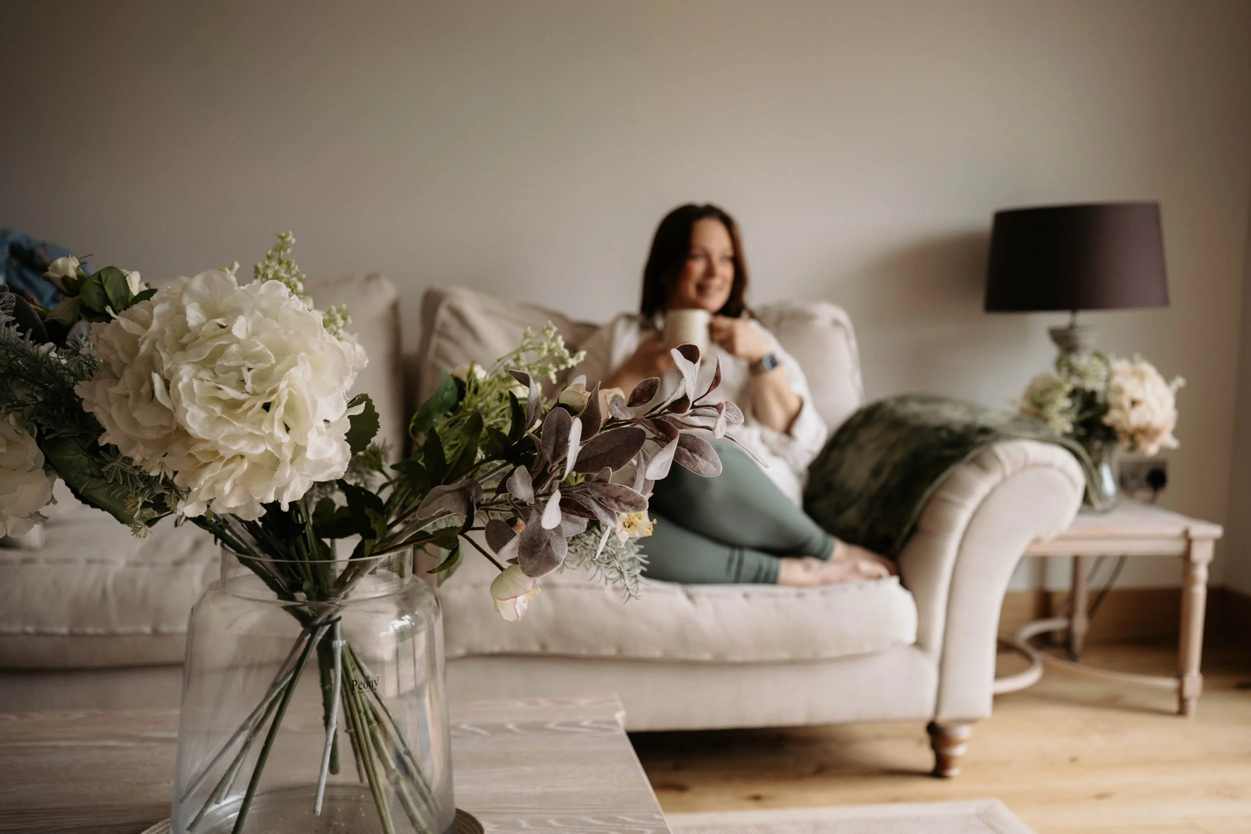 A woman sitting on a beige sofa in a living room, smiling and holding a mug, with a vase of white and purple flowers in the foreground.