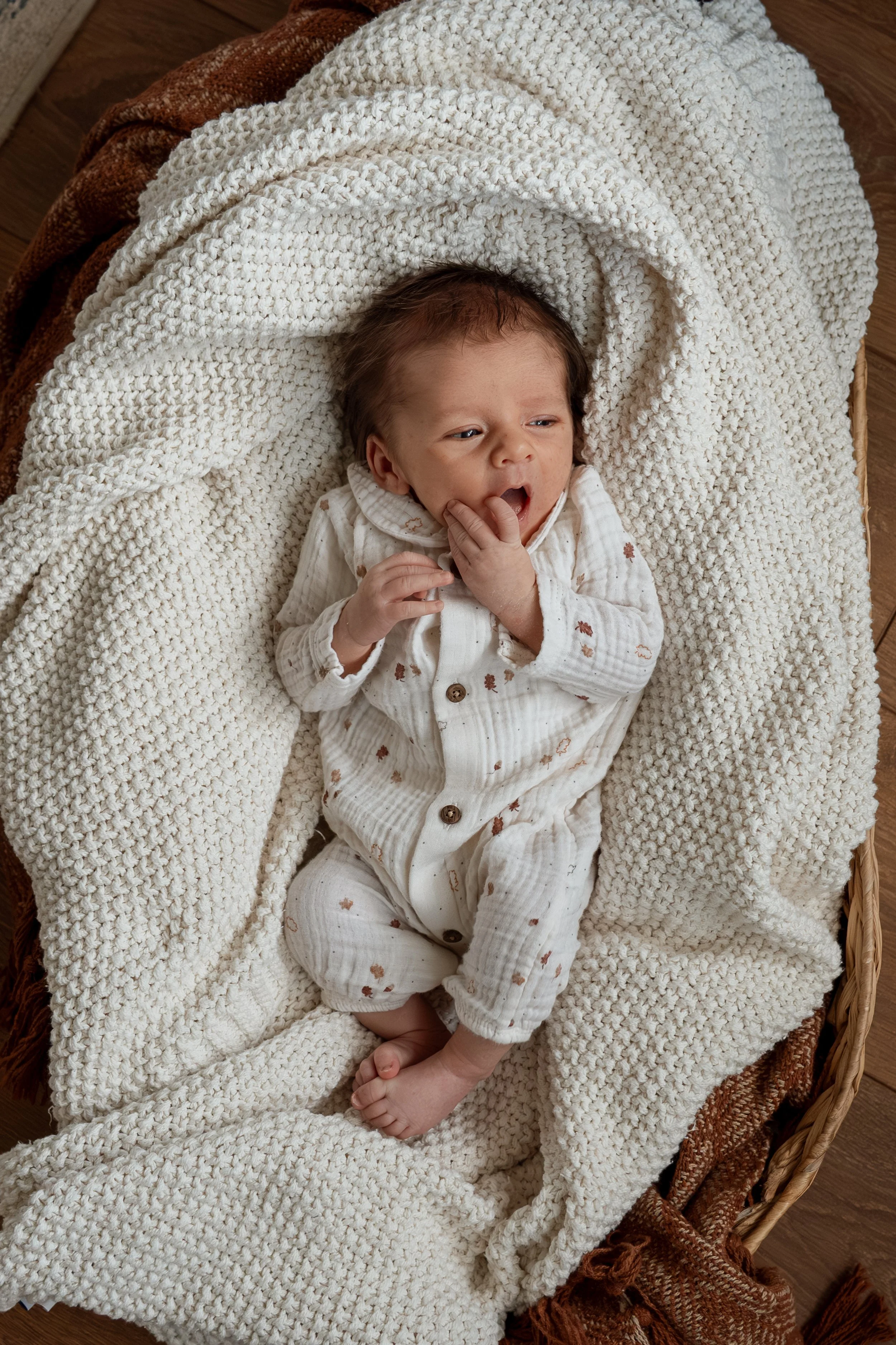 A baby lying on a cozy, cream-colored knitted blanket in a basket, wearing a white outfit with small brown patterns, yawning or sighing with fingers in mouth.