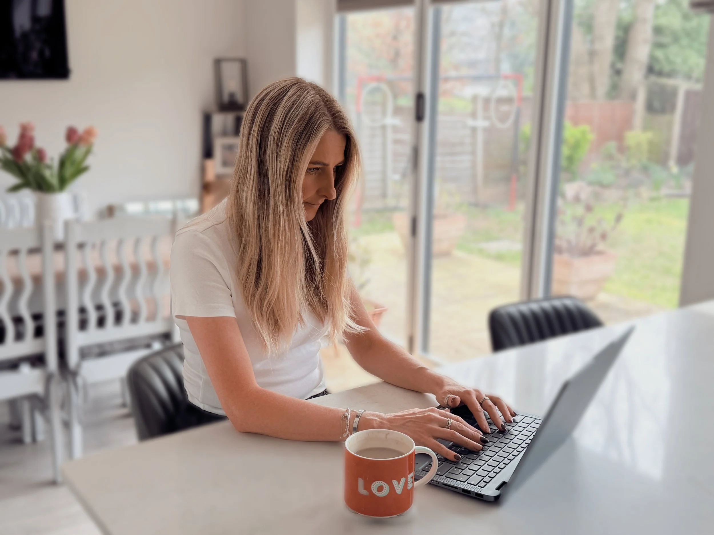 A woman with long blonde hair, wearing a white t-shirt, sitting at a kitchen table using a laptop. There is a mug with the word 'LOVE' on it in front of her and a flower vase with pink flowers on a white chair in the background. Large glass sliding doors reveal a garden outside.