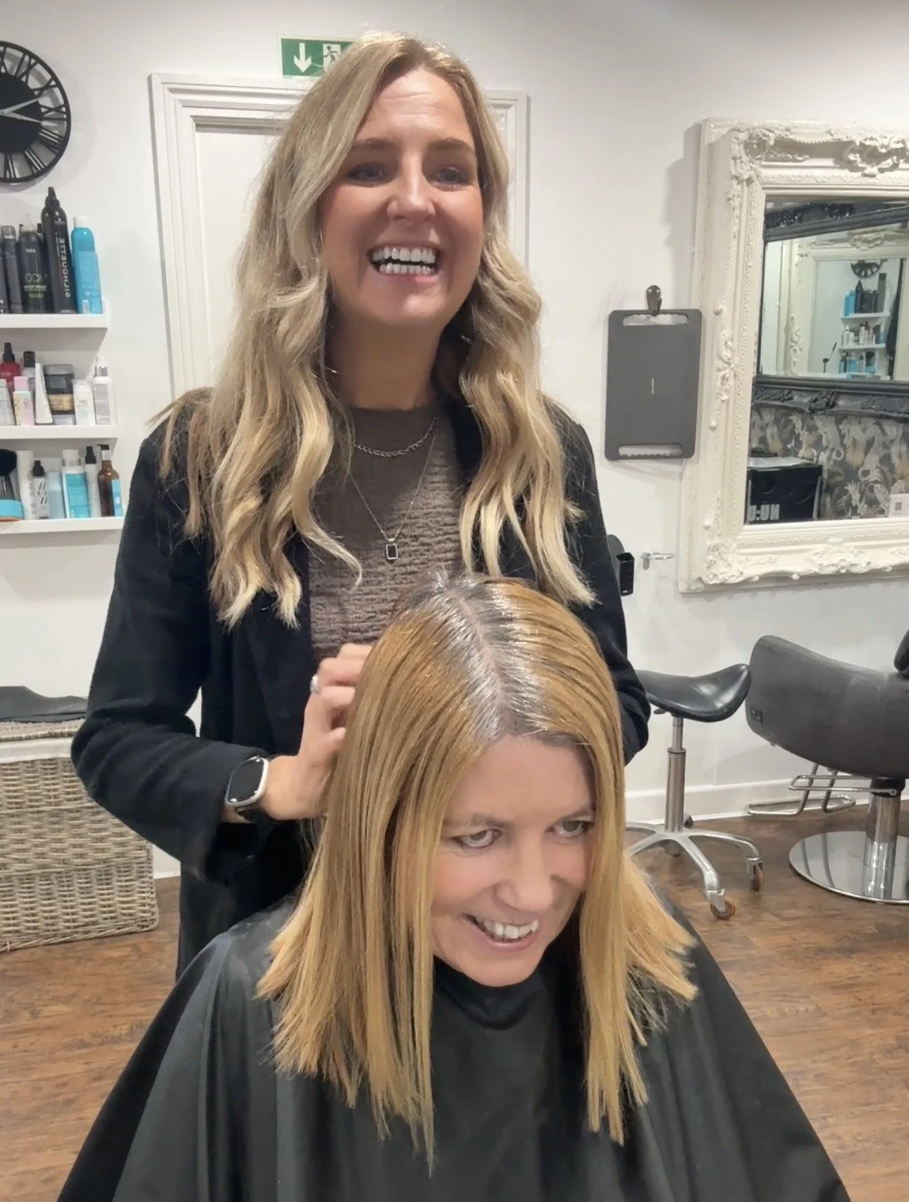 Woman with long, blonde hair getting her hair dyed in a salon. She is seated with a black cape, and another woman stands behind her smiling.