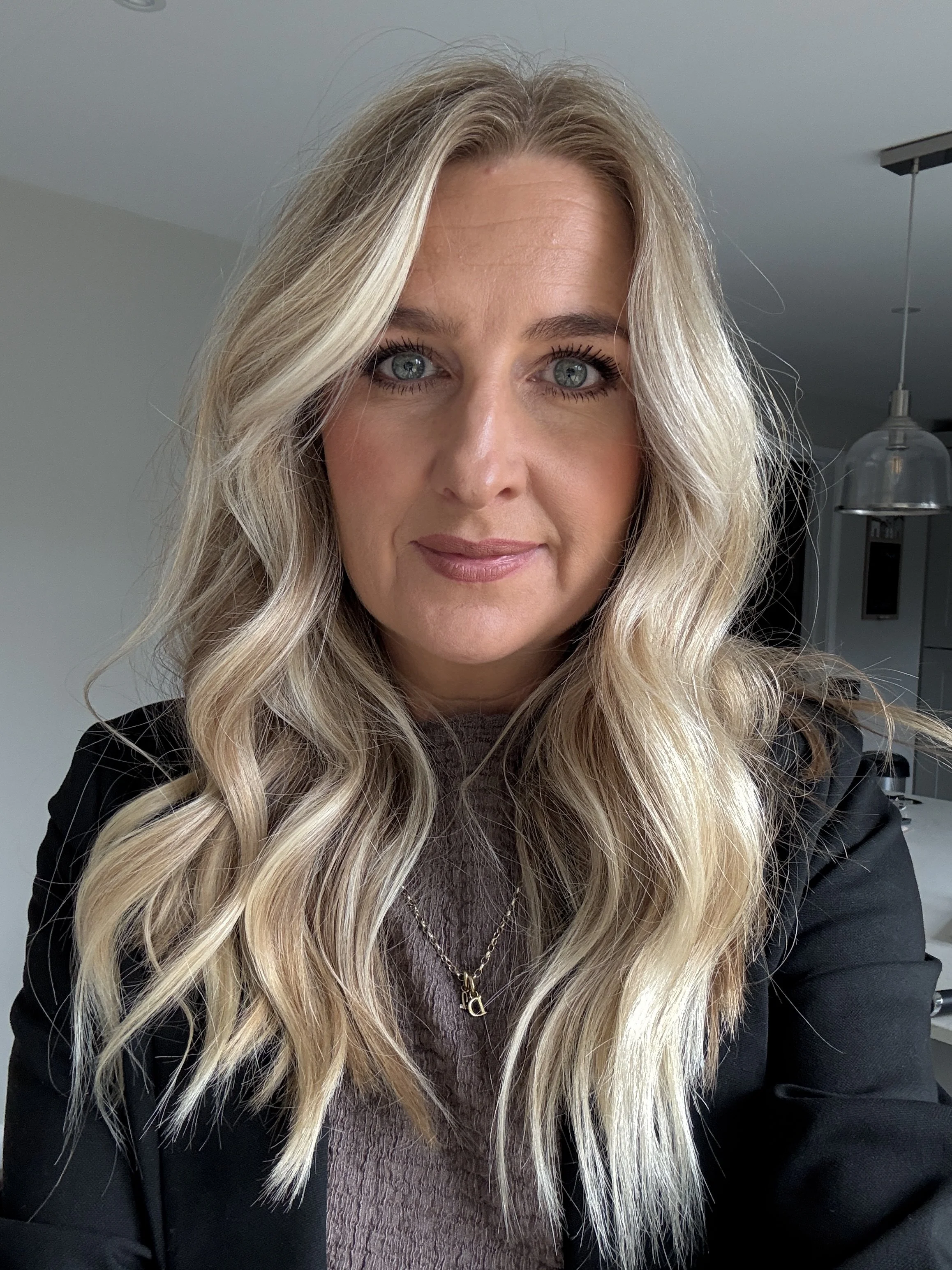 A woman with long, wavy blonde hair and blue eyes is taking a selfie indoors. She is wearing a black blazer and a gray top, accessorized with a gold necklace featuring a pendant. The background shows a modern kitchen with a pendant light and some cabinets.