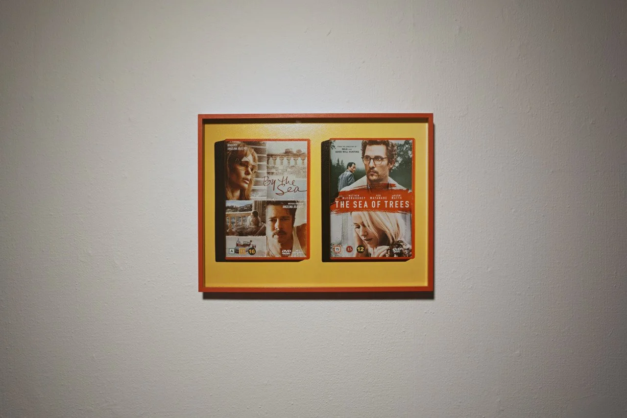 Framed display of two DVD cases, one titled 'By the Sea' and the other 'The Sea of Trees', mounted on a plain wall.