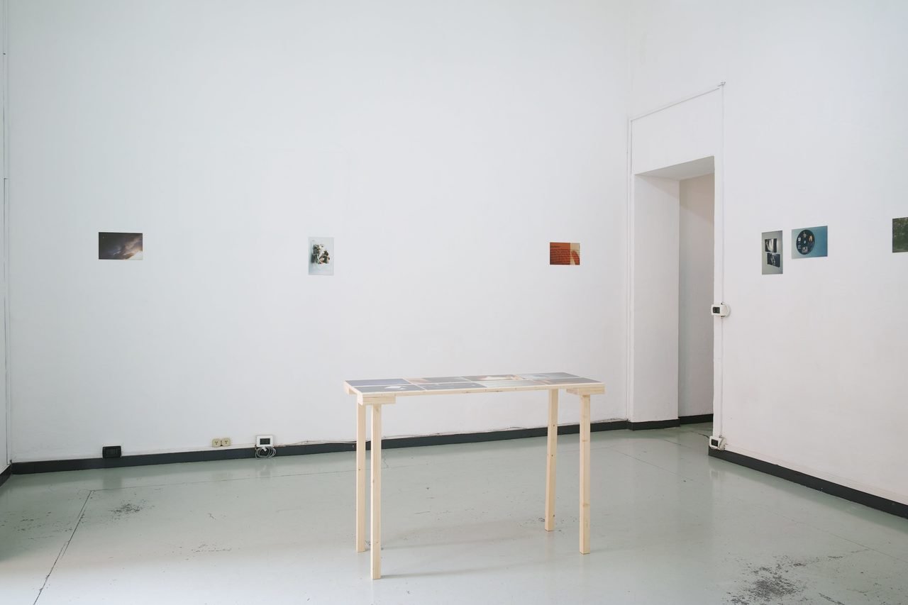 An art gallery with white walls, small framed photos on the walls, a light wooden table with papers or photographs, and some electrical outlets and wires on the floor. Space 4235, Genova.