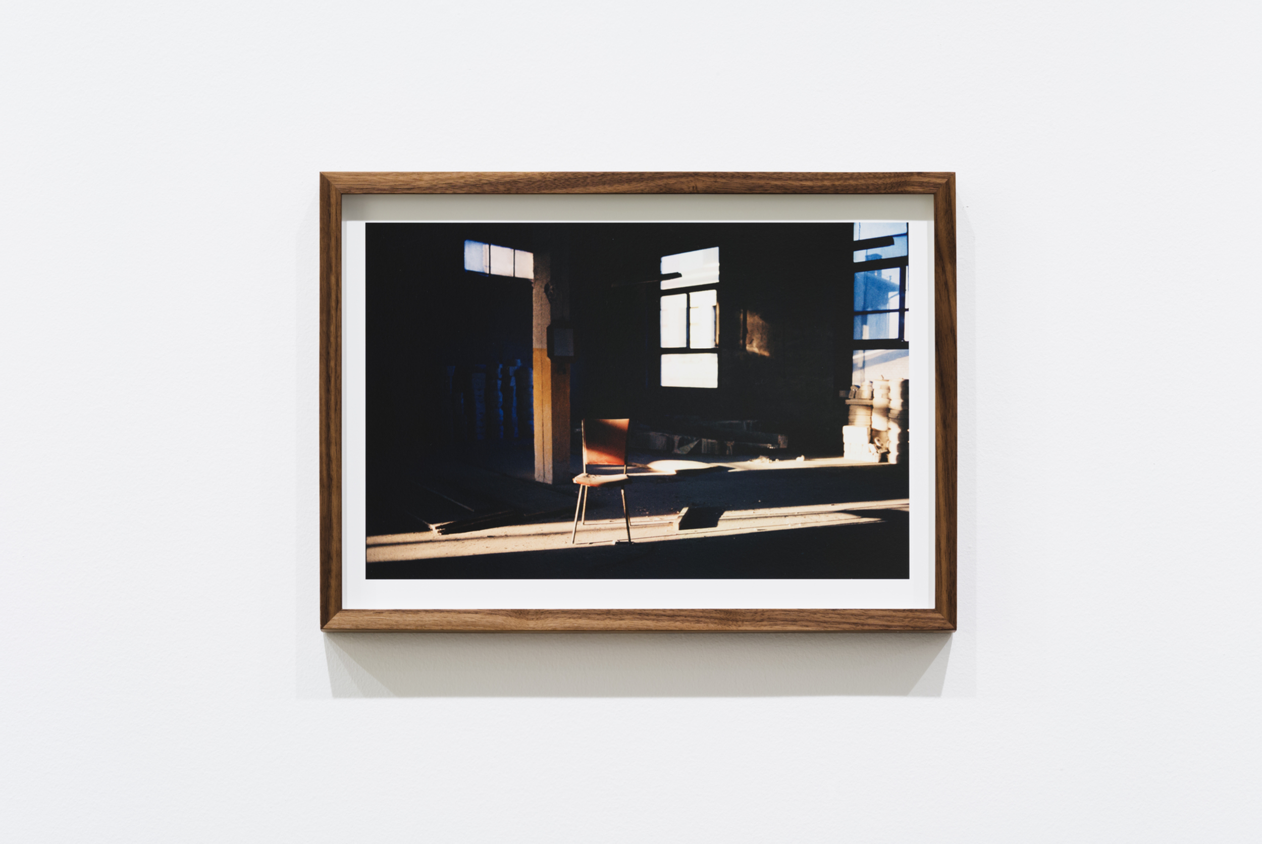 A framed photograph of an interior space illuminated by natural light from windows, with a wooden chair placed in the center.