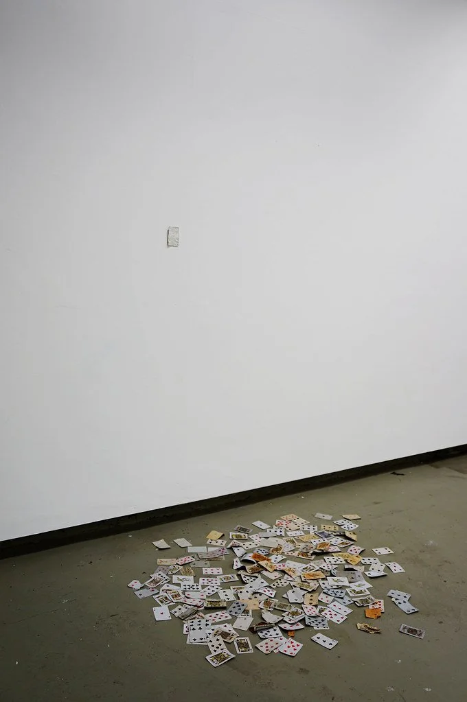 A scattered pile of playing cards on a bare, gray floor against a white wall.