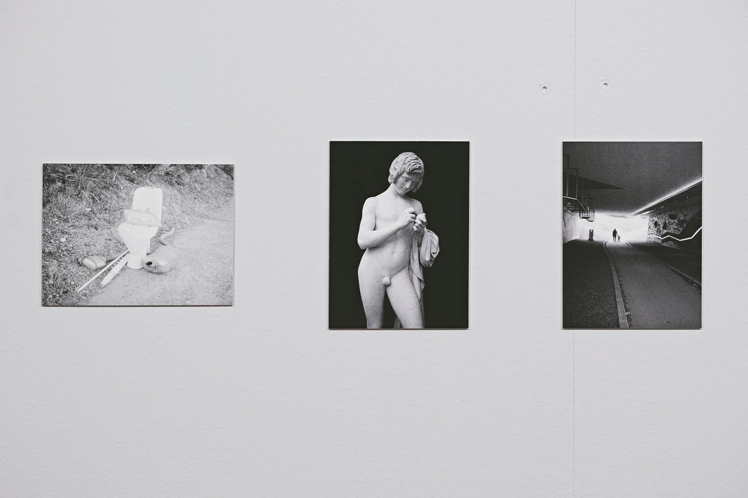 Three black and white photographs displayed on a white gallery wall. The first photo shows a discarded toilet with items around it on a sidewalk. The second photo depicts a classical statue of a young girl with an object in her hand, against a dark b