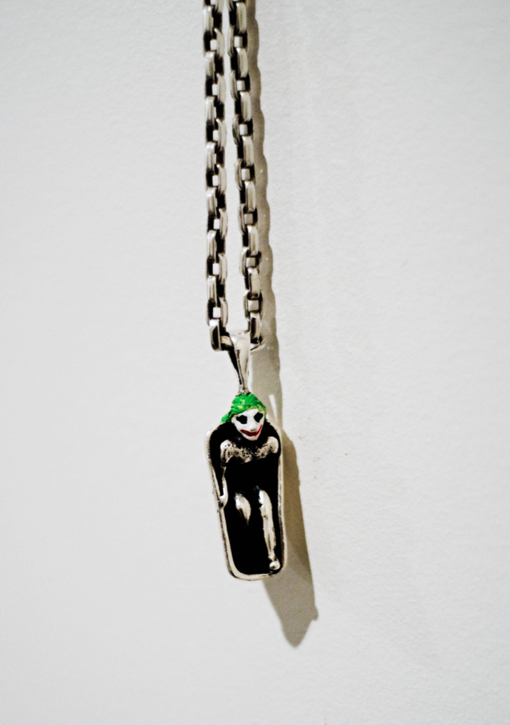 A silver necklace with a pendant of a cartoon Joker character wearing a green hat, with makeup and a grim expression, hanging against a plain white background.