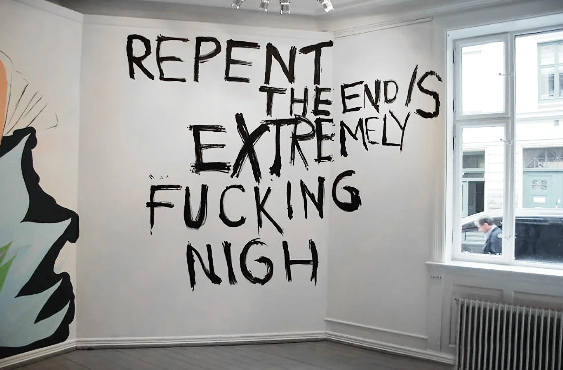 Wall art with the words "REPENT THE END IS EXTREMELY FUCKING NIGHT" written in bold black letters inside a room with a window showing a street scene outside.