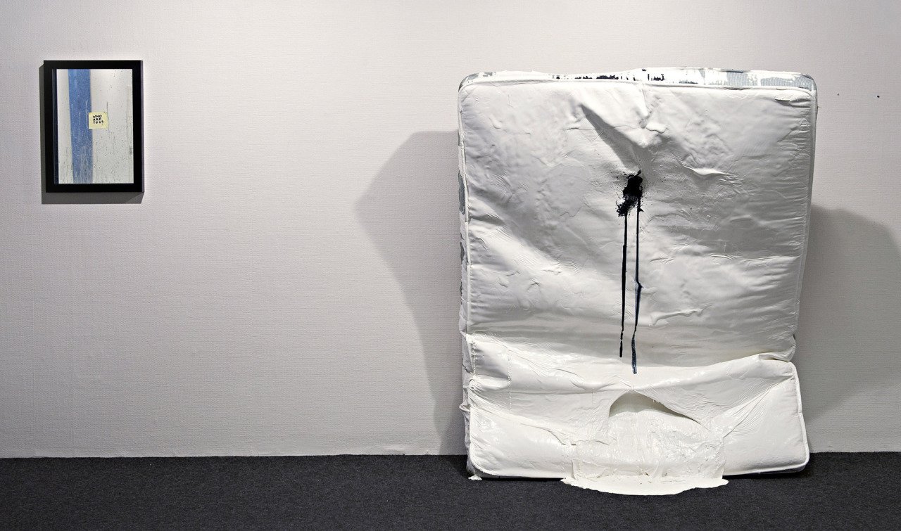 A white mattress leaning against a white wall with a black spot and black drips on it, next to a small framed art piece on the wall with a note saying WHO ARE YOU?