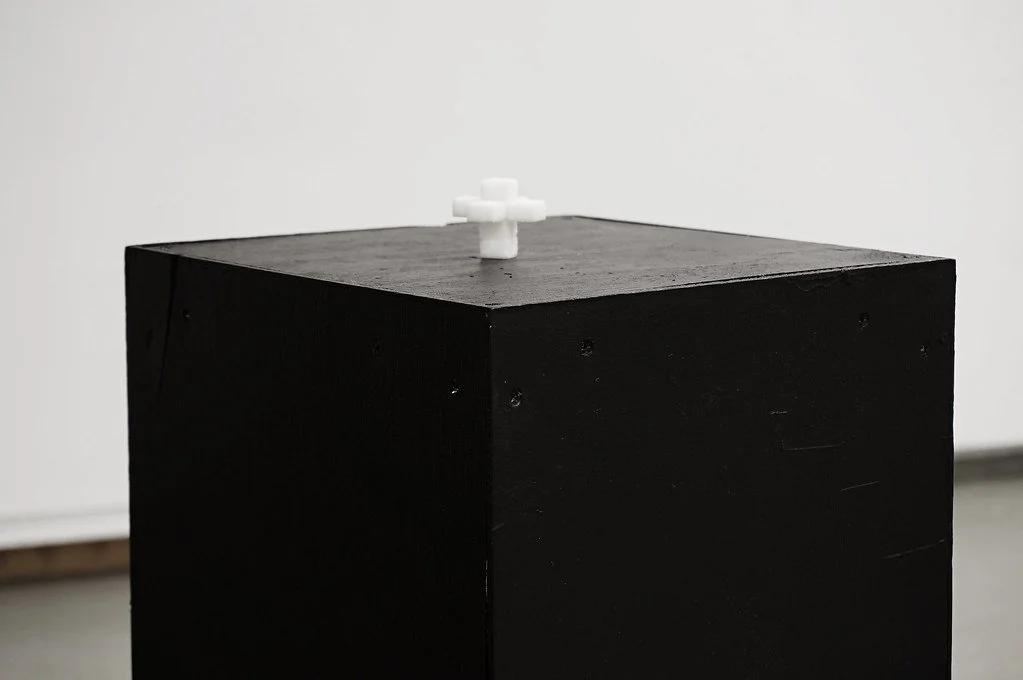 A black pedestal with a small white sculpture of a cross on top, placed against a plain white background.
