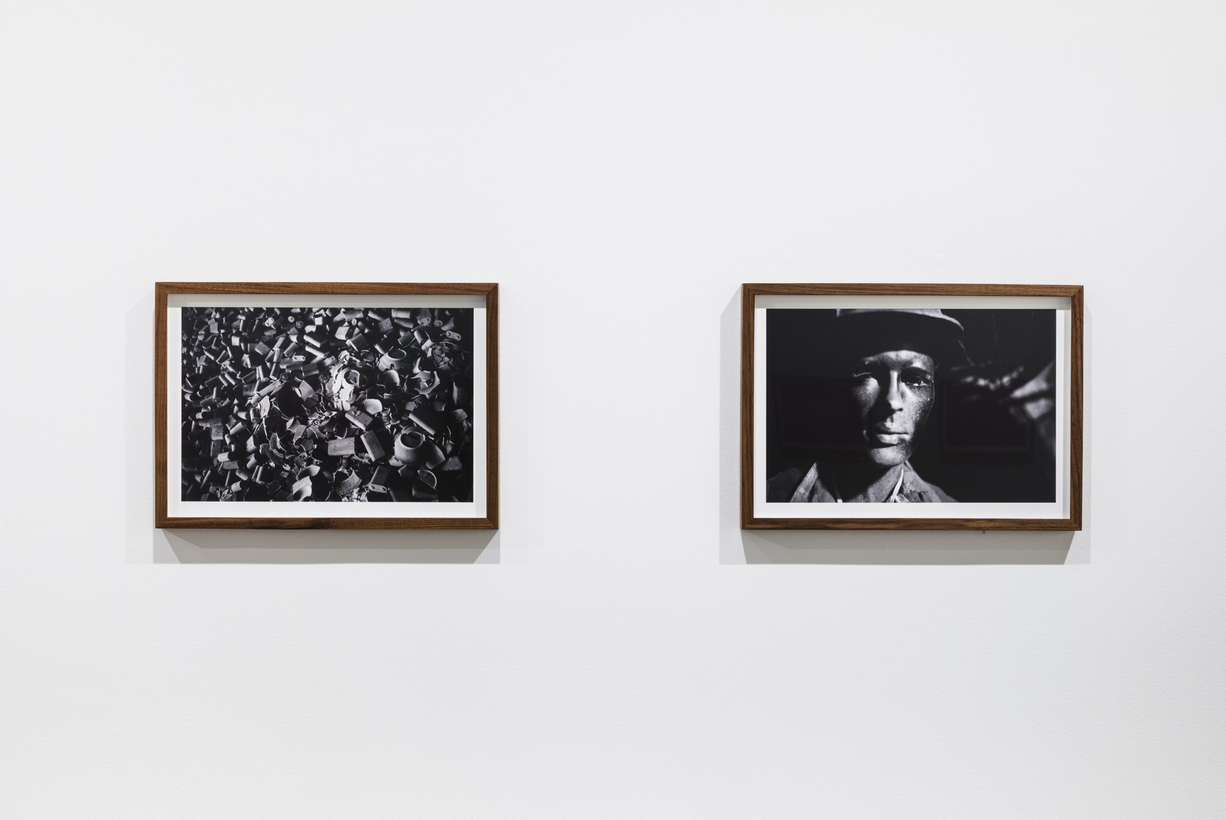Two black and white photographs framed in wood hung on a white gallery wall. The left photo depicts a chaotic pile of small, broken objects, while the right photo shows a woman's face with a serious expression, wearing a wide-brimmed hat.