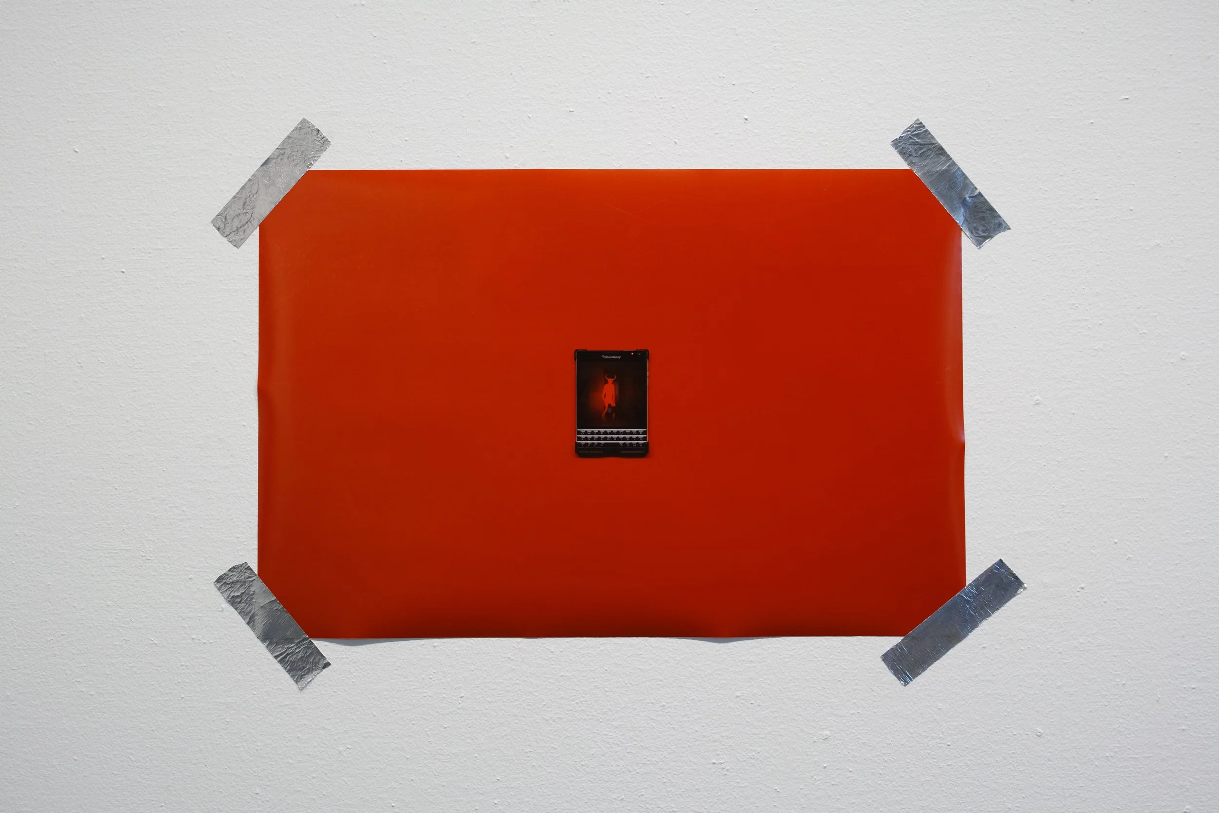 A BlackBerry smartphone displaying a devil on its screen, mounted on a red background taped to a white wall with four pieces of silver duct tape.