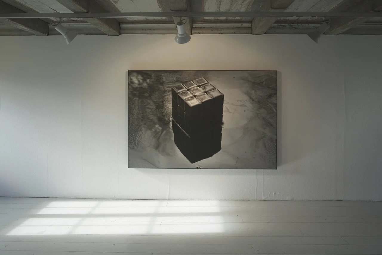 Black-and-white artwork of a stack of closed books on top of a floating cube, reflected in a puddle of water, displayed on a gallery wall.
