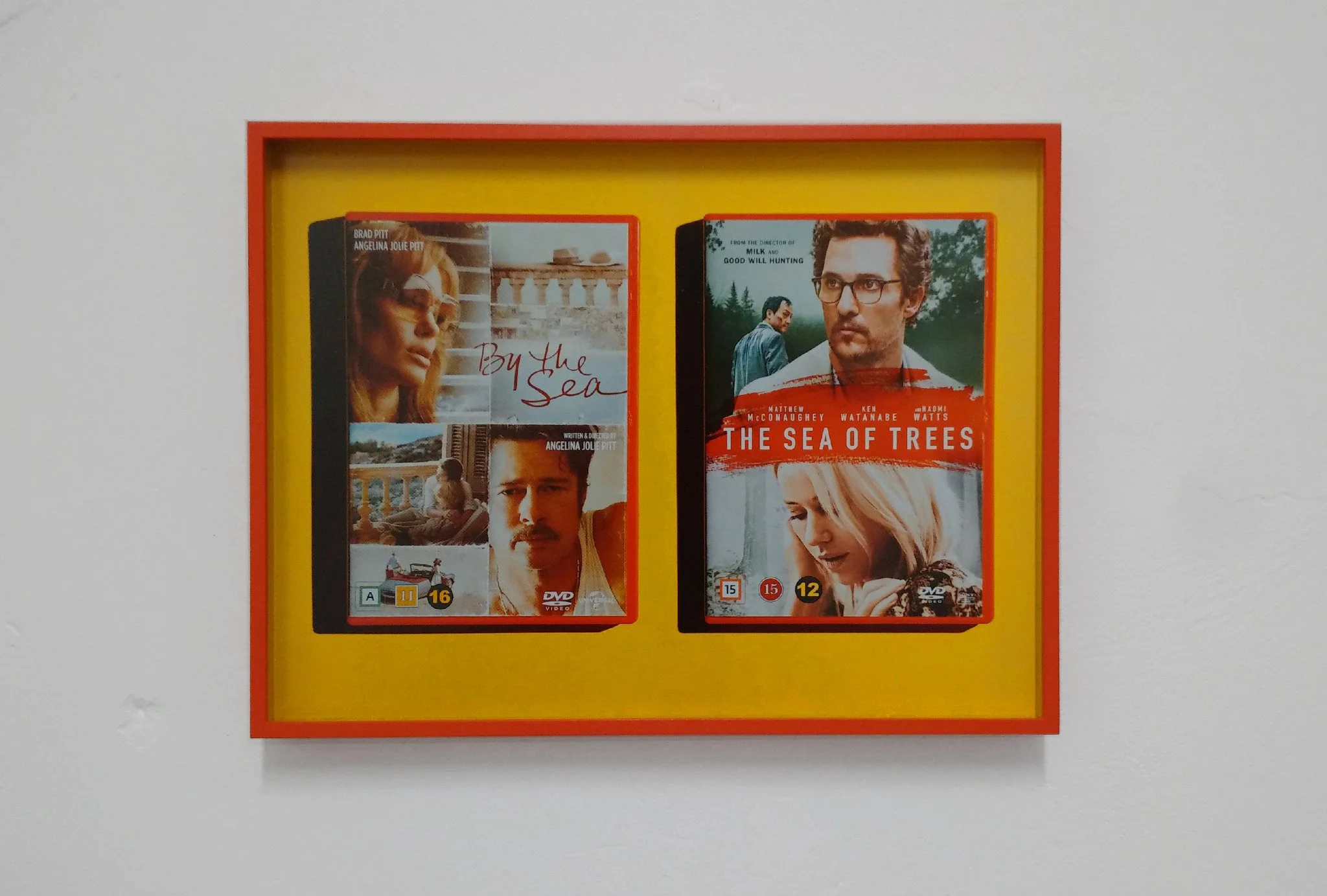Two DVD cases, one titled 'By the Sea' with Angelina Jolie and Brad Pitt, and the other titled 'The Sea of Trees' with Matthew McConaughey and Naomi Watts, displayed inside a red and yellow frame on a white wall.