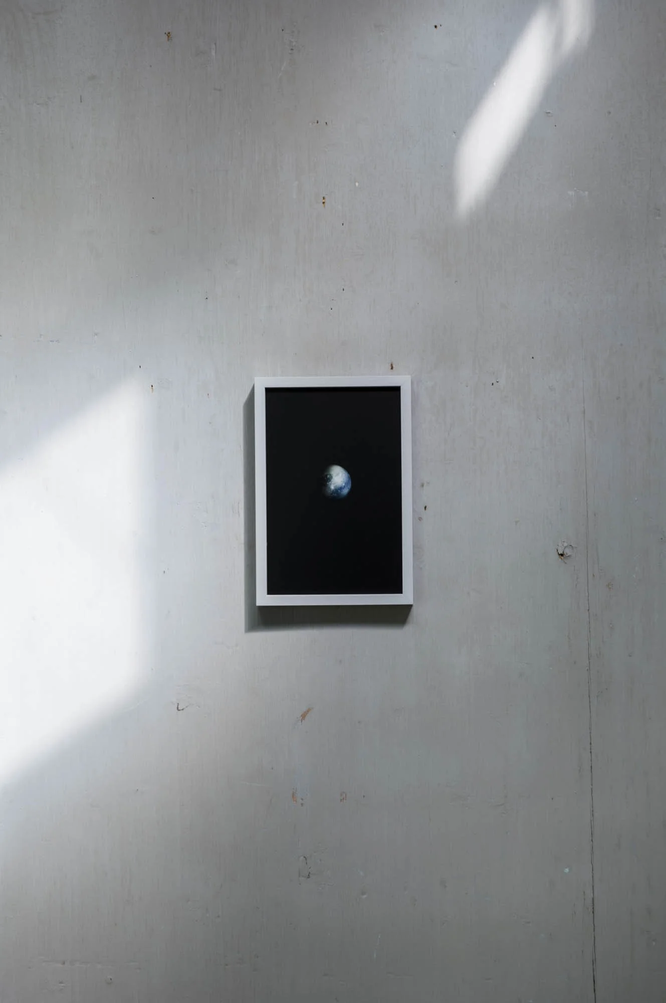 Photograph of a framed picture of Earth hanging on a plain wall with soft natural light and shadows.