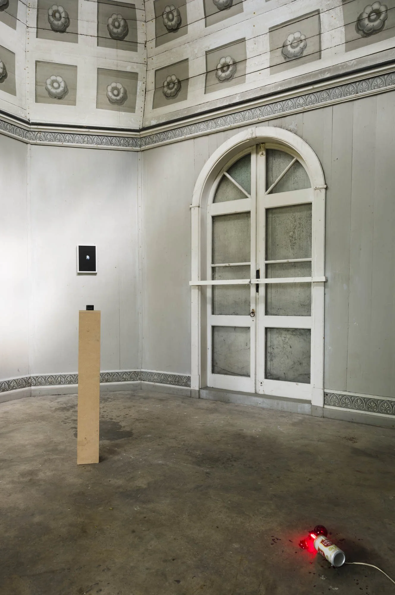 Empty room with ornate ceiling, arched window with frosted glass, a small black screen on the wall, a pedestal with a device, and a fallen bottle with a red light on the floor.