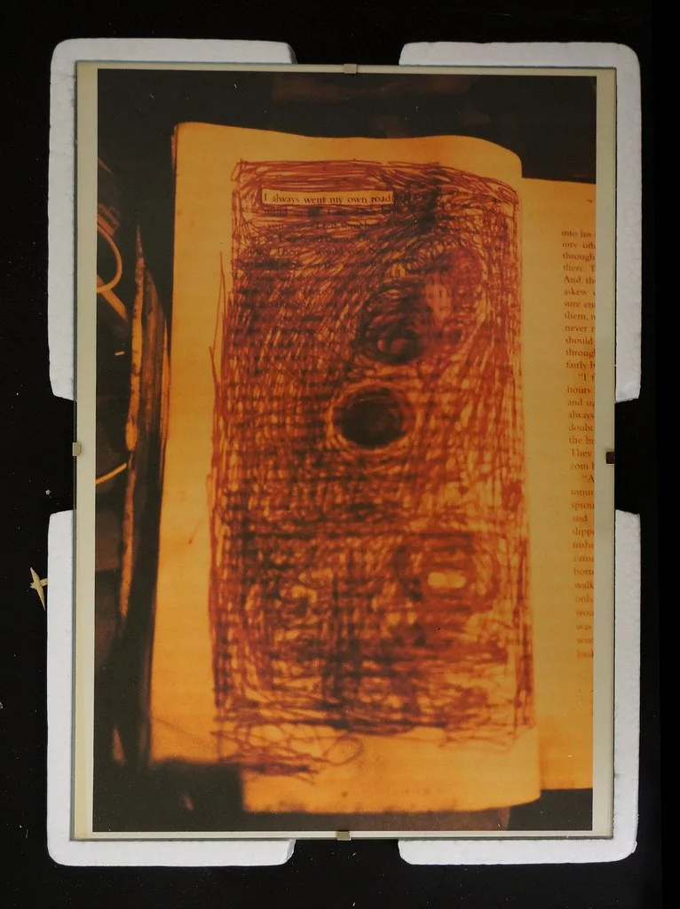 A page from a book with a red and orange scribbled drawing over it, partially obscuring the text. The drawing appears to be made with a pen or marker and covers most of the visible text.