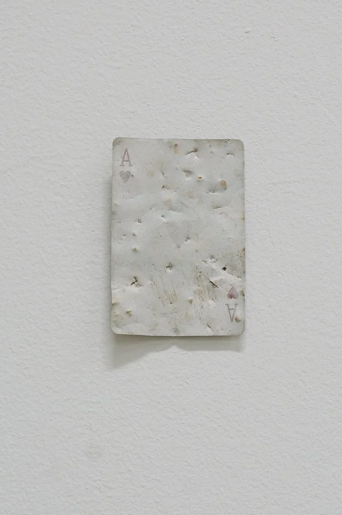 An old, weathered playing card, the Ace of Hearts, mounted on a white wall.