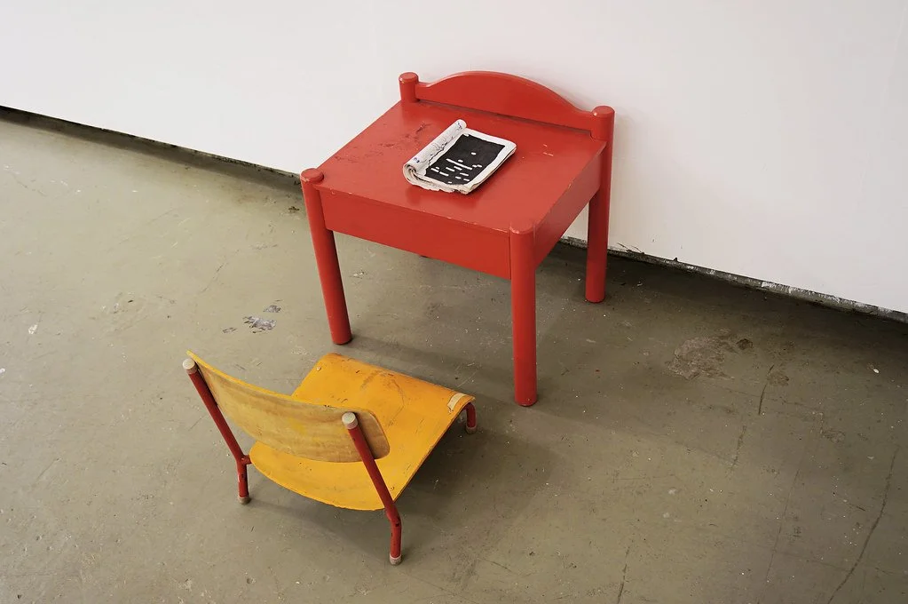 A small red table with a black and white object on top, next to a yellow knee-high chair with red legs, on a concrete floor in front of a white wall.