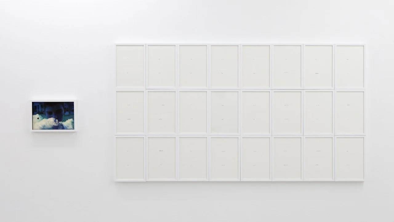 An art installation featuring a large white grid with empty frames and a small framed photo of a person behind clouds on a white wall.