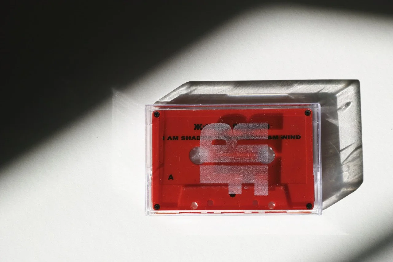 A red cassette tape inside a clear plastic case, casting a shadow on a white surface. The album I am Shadow I am Ghost I am Wind. Frøya Records.