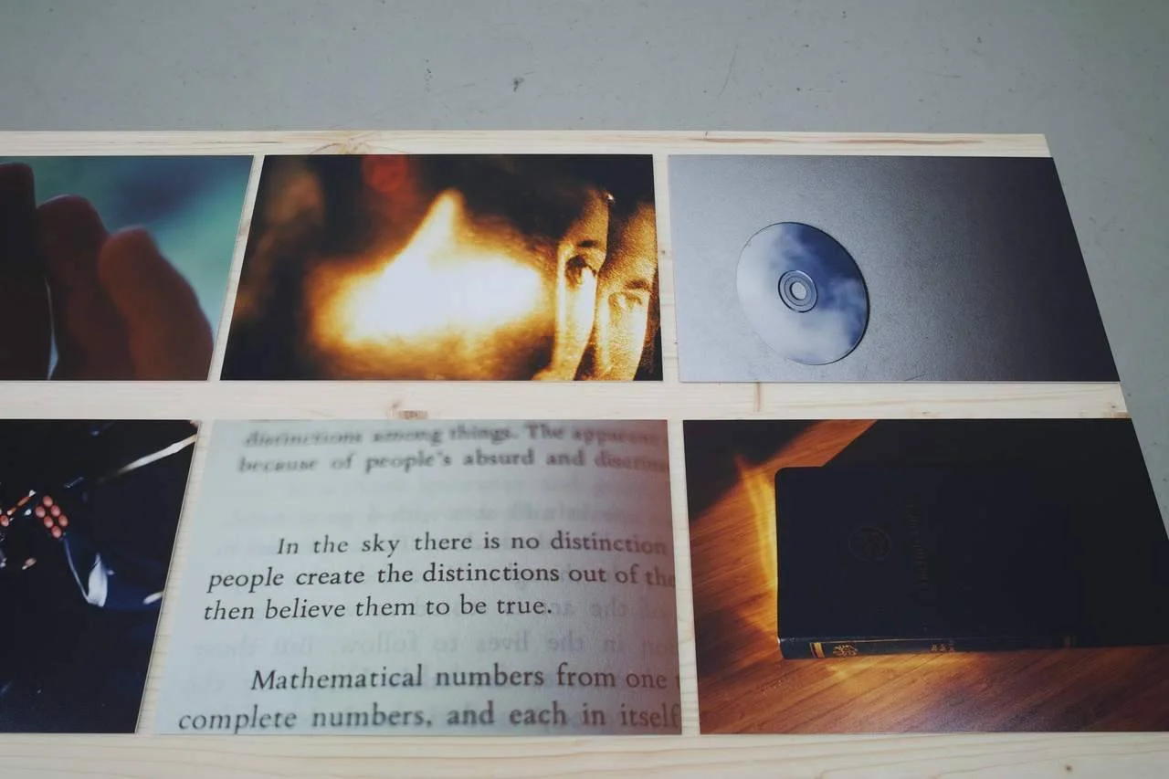Photograph of a wooden frame with several printed images and a CD placed on a gray surface. The printed images include a blurry photo of a person, a close-up of a book page with text including 'In the sky there is no distinction', and a photo of a bl