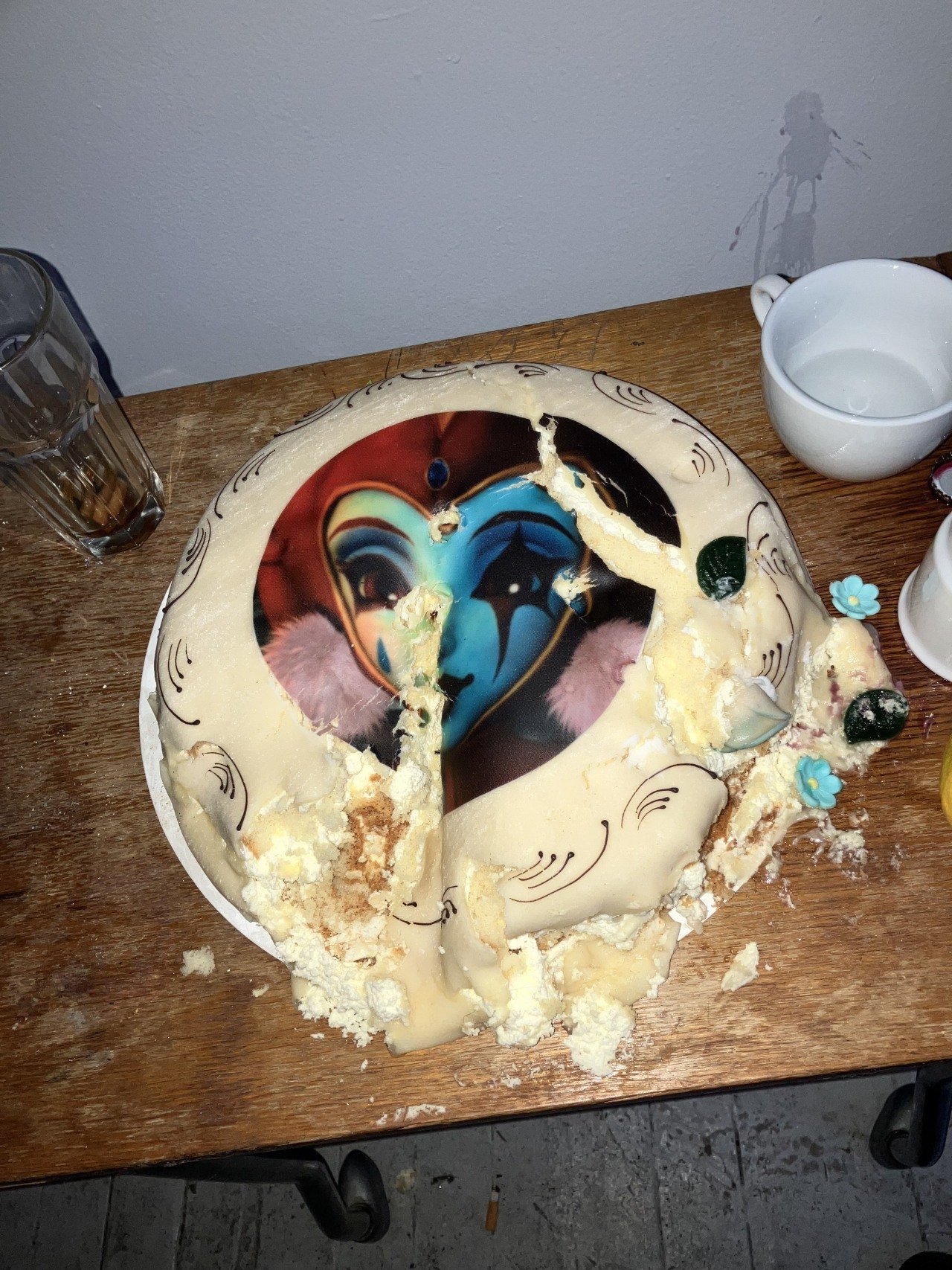 A partially eaten birthday cake with a harlequin face image on top, placed on a wooden table with a glass of water, a white mug, and some decorations around.