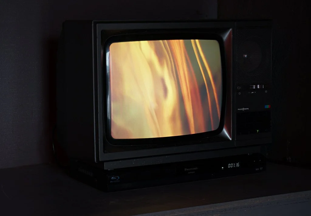 A vintage CRT television displaying a colorful abstract image, placed on top of a black device in a dark room.