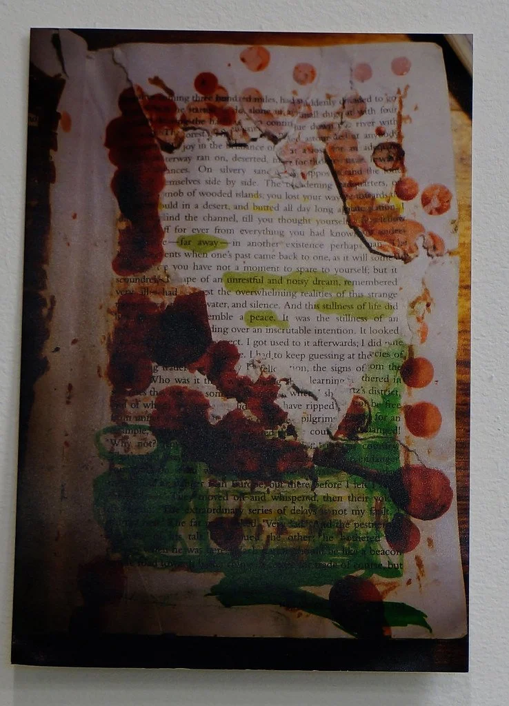 A mixed media artwork on paper with ink and paint, featuring a person's face outline made with dark, abstract strokes, placed over a background of printed text. The face has red and orange paint accents, and there are yellow highlights on the text ar