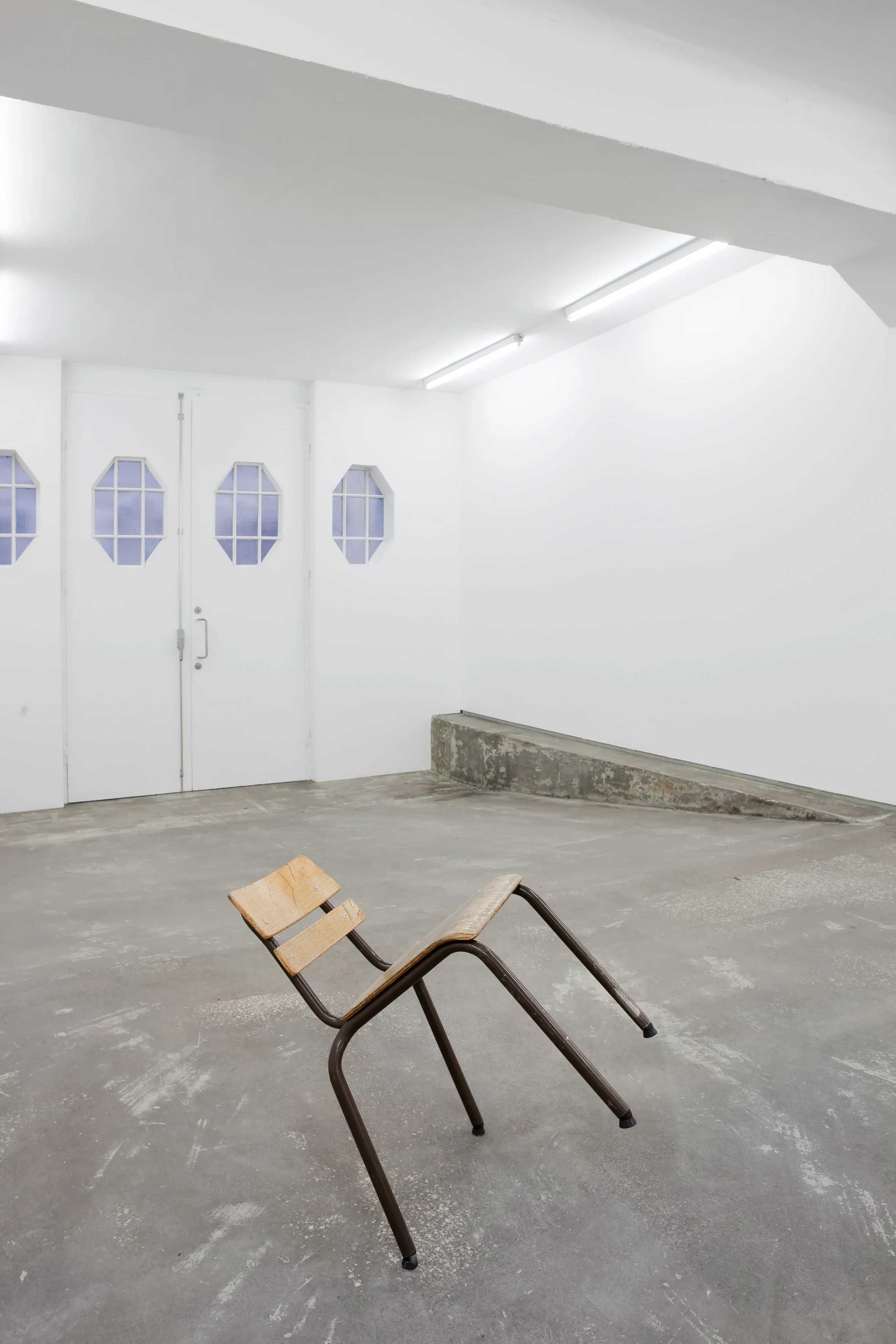An empty basement with a tilted wooden chair on the concrete floor and white walls and ceiling, illuminated by fluorescent lighting.