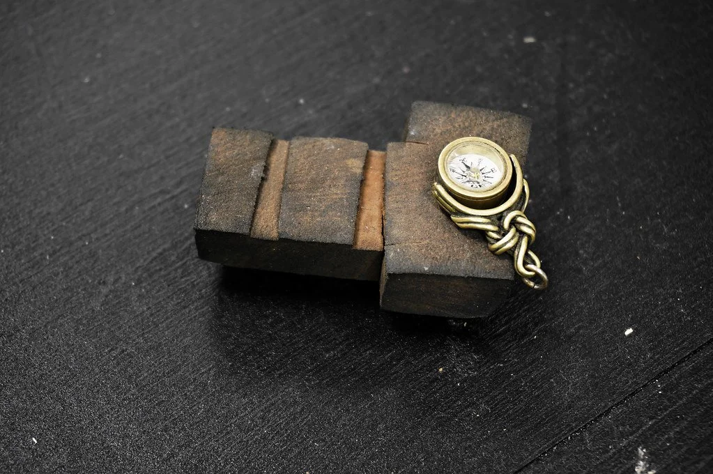 A small brass compass attached to a metal chain, resting on a dark, rough-textured wooden block.