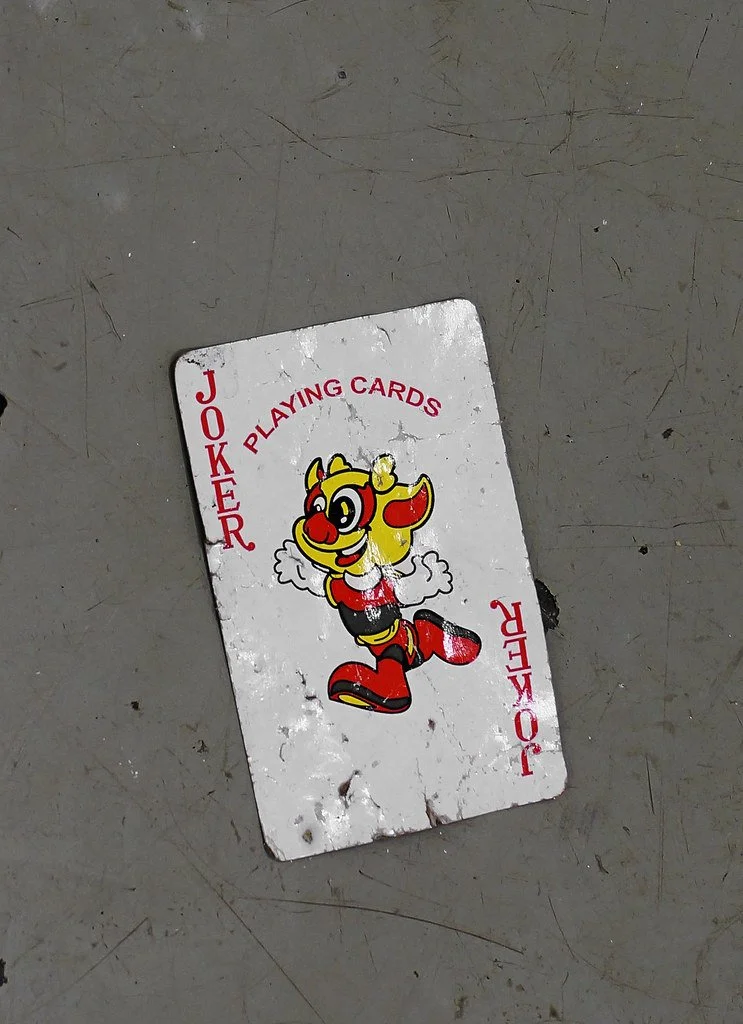 A worn playing card lying on a gray surface, featuring a cartoon character with yellow hair, red boots, and a black outfit, with the words 'JOKER' and 'PLAYING CARDS' printed on it.
