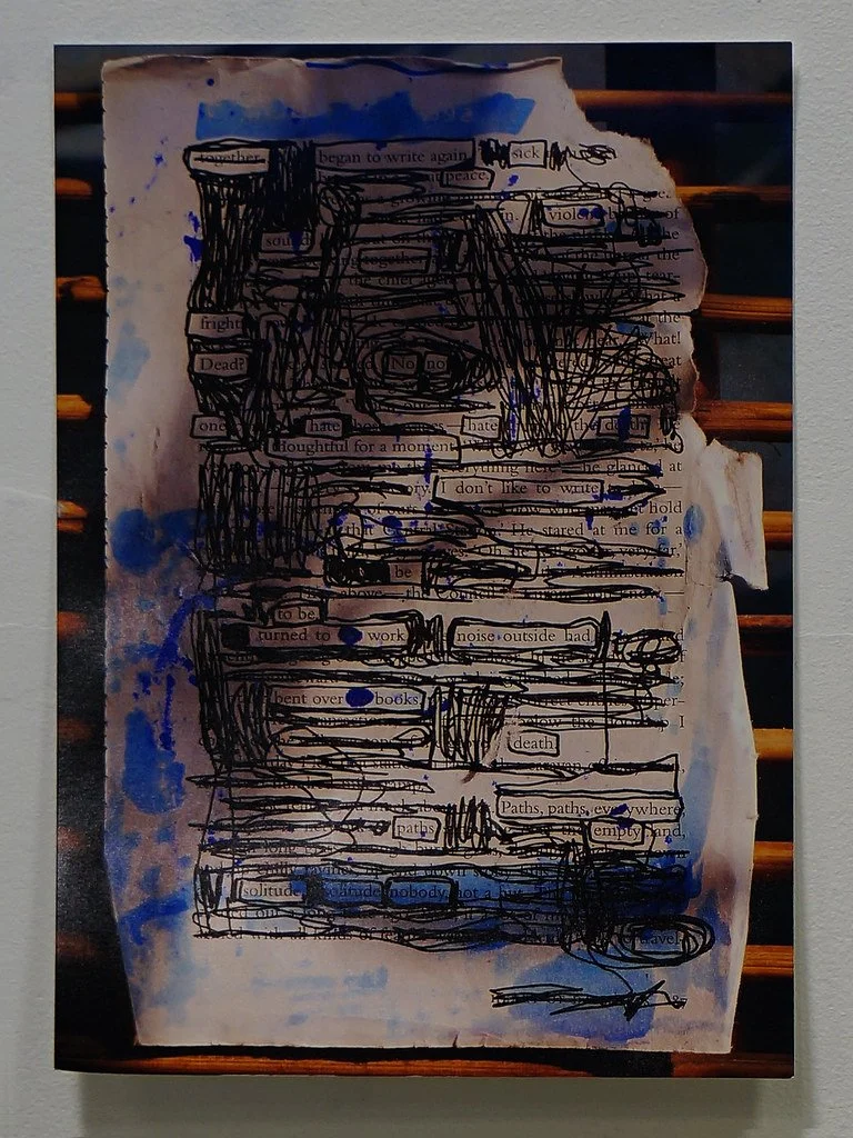 A piece of paper with handwritten and scribbled black ink over typed text, some areas painted with blue watercolor, placed on a wooden surface. The paper is torn at the top right corner.