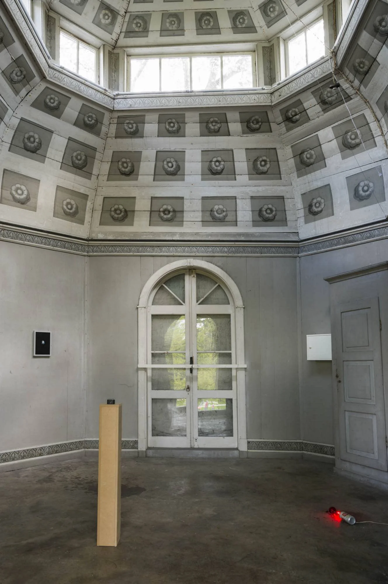 Empty room with high ceiling and patterned wall panels, a large window at the top, a closed arched window/door at the bottom, a wooden pedestal with an electronic device, and a small red laser pointer on the floor.