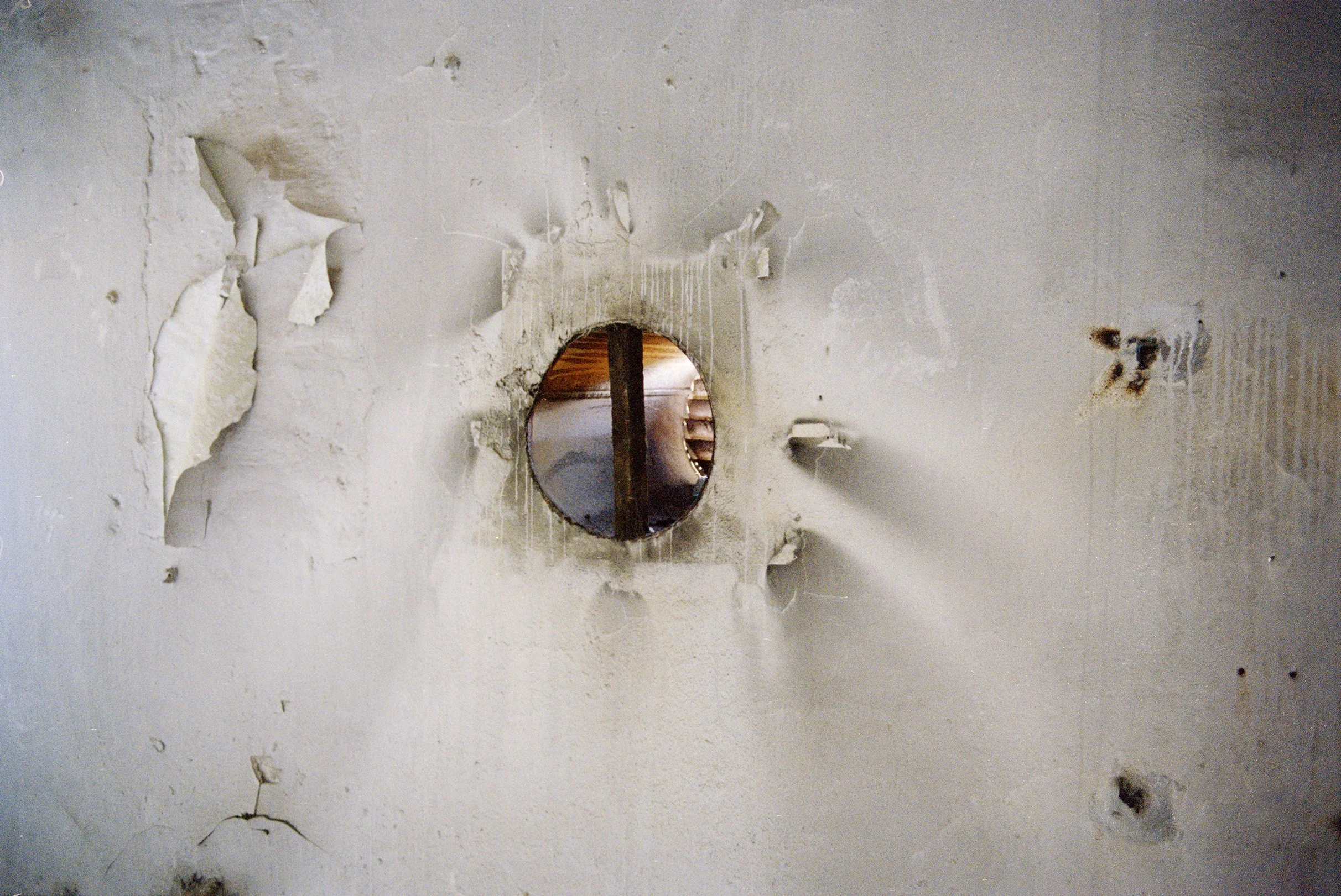 A hole in a white, cracked wall with a mirror or reflective surface behind it, revealing a staircase or wooden structure.