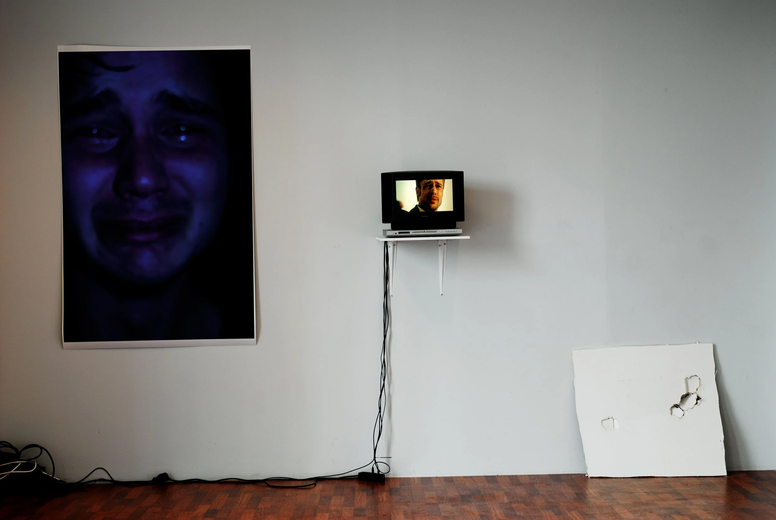 An art installation includes a large portrait of a person's face with a sad expression on the left, a small television screen showing a man's face in the center, and a white canvas with holes on the right against a plain wall.