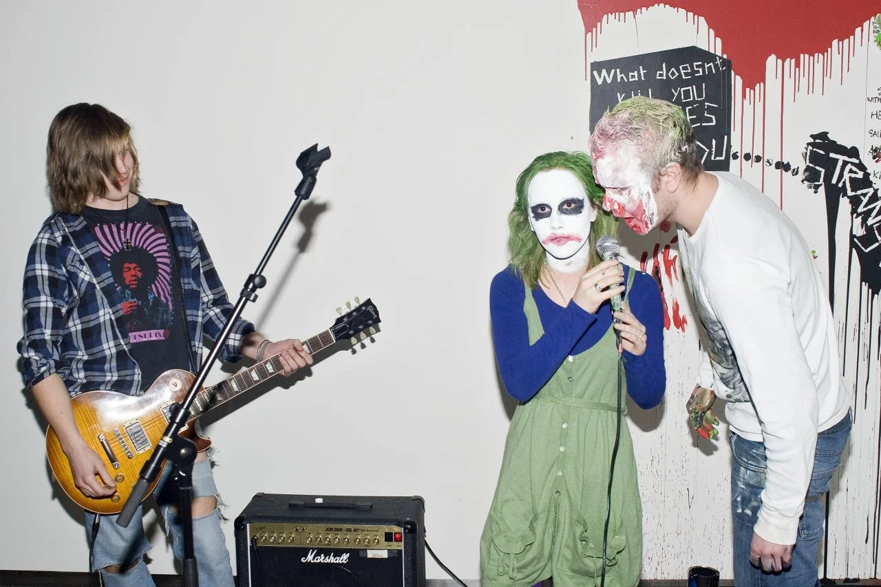 A woman with clown makeup and green hair sings into a microphone while a man with clown makeup leans toward her, holding a microphone. A person with long hair and a plaid shirt plays an electric guitar nearby. The scene appears to be a performance or