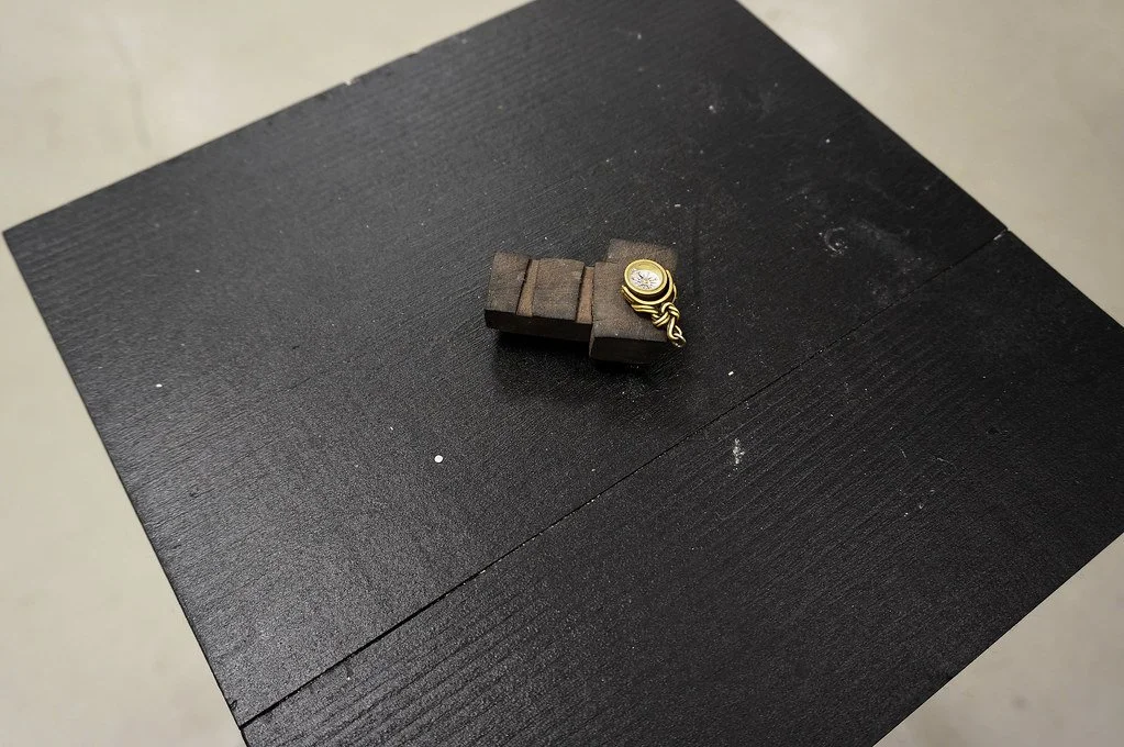 A small, round gold locket with a chain and a clear gemstone, resting on a piece of wooden jewelry display on a black textured surface.