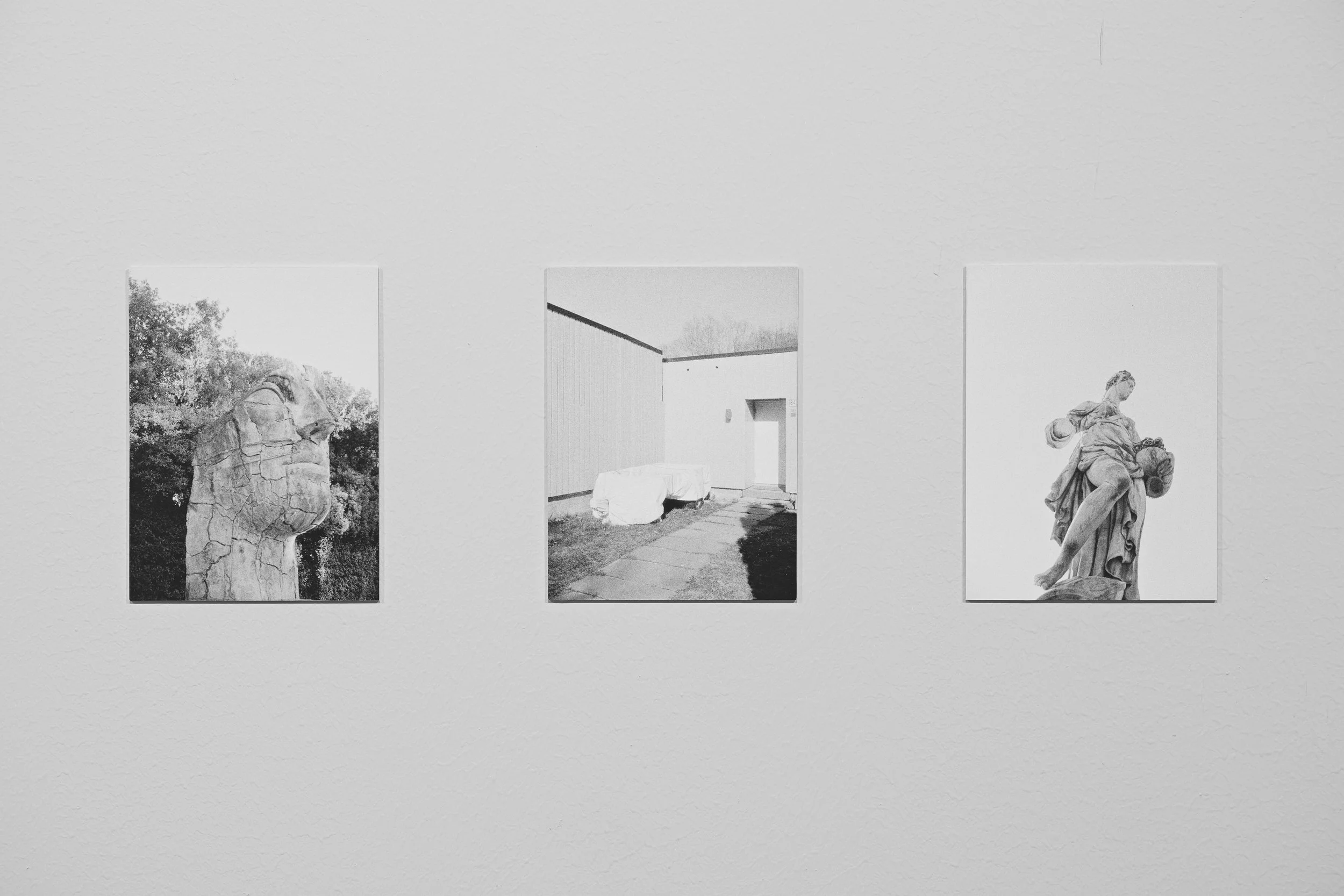 Three black and white photographs displayed on a white wall. The first photo shows a cracked stone sculpture of a human face, the second features a minimalist outdoor scene with a covered object and a building, and the third depicts a classical statu